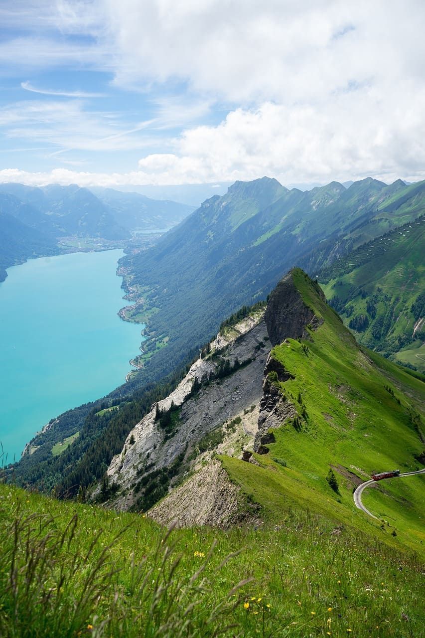 Vue en plongée d'une crête montagneuse verdoyante près d'un lac turquoise, avec un petit train rouge circulant sur une voie.