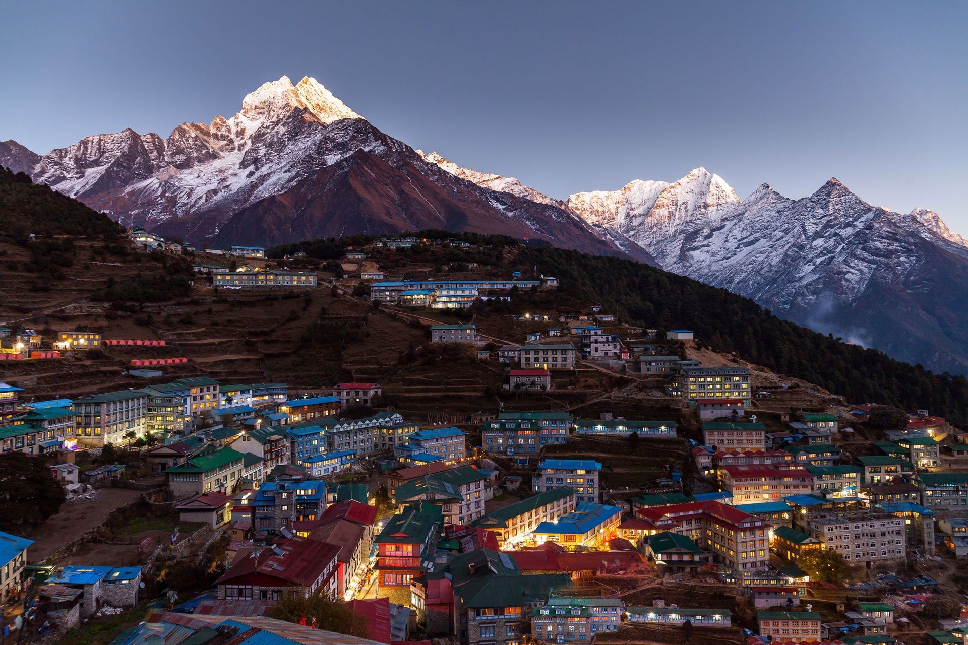 Un villaggio illuminato su un pendio terrazzato al crepuscolo, con grandi cime montuose innevate sullo sfondo.