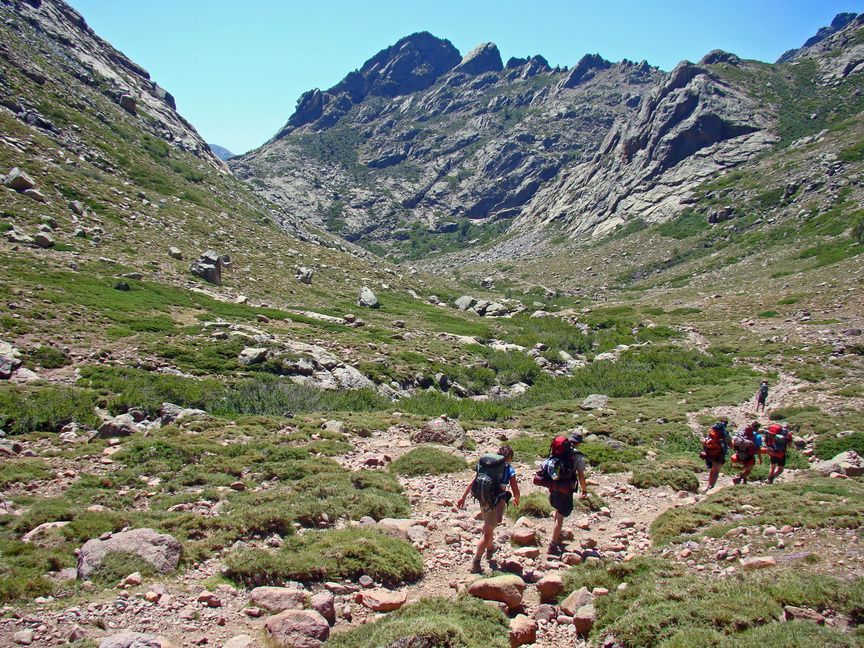 Un groupe WeRoad en randonnée avec de gros sacs à dos sur un sentier rocheux dans une vaste vallée de montagne.
