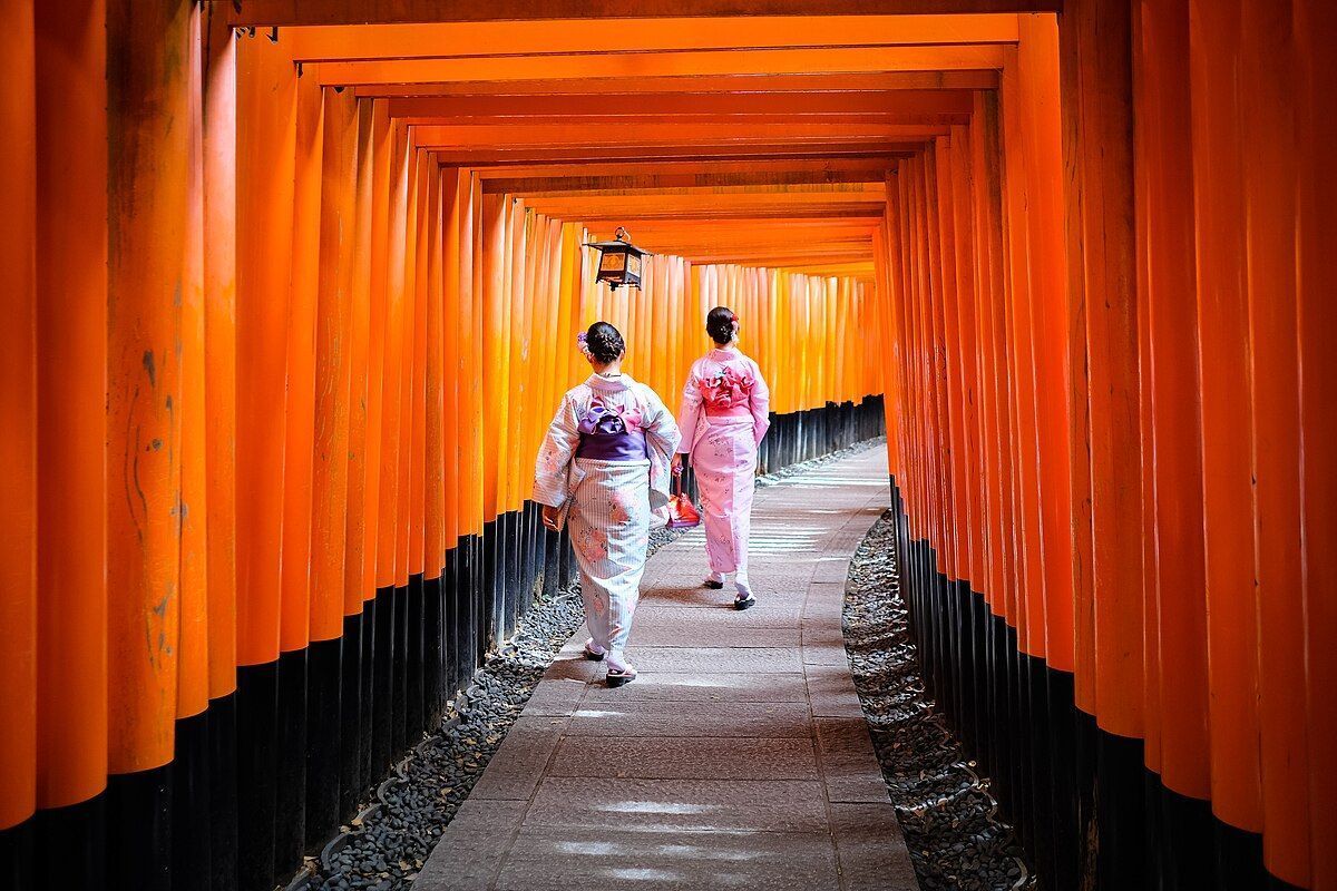 Due donne in kimono tradizionali camminano lungo un sentiero di pietra attraverso un tunnel formato da portali arancione brillante.