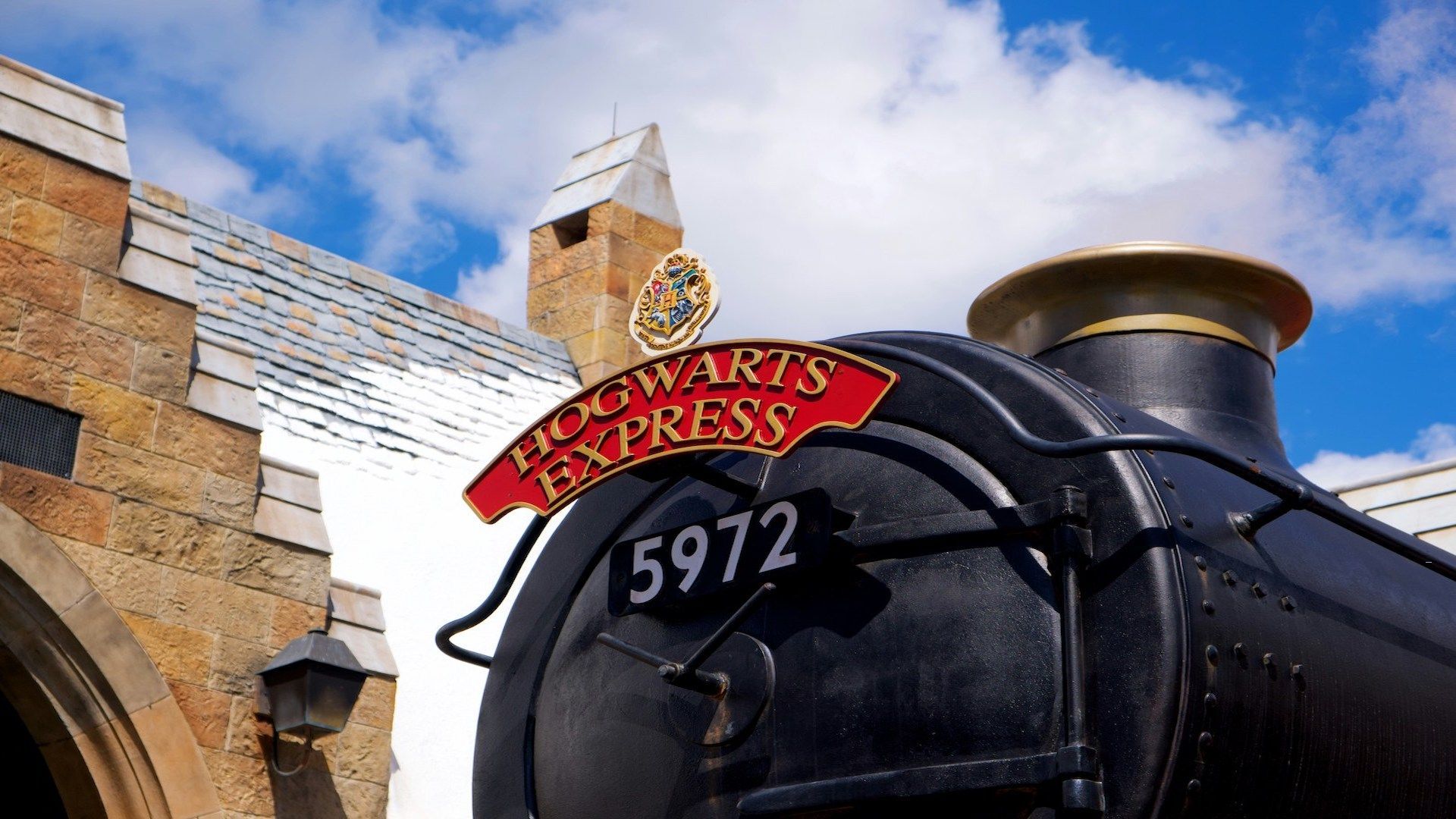 Primo piano dal basso del treno a vapore Hogwarts Express, numero 5972, con un edificio in pietra sullo sfondo.