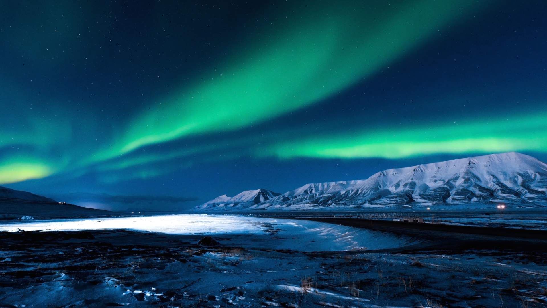 L'aurora boreale verde risplende sulle montagne innevate e su un paesaggio ghiacciato sotto un cielo notturno stellato.