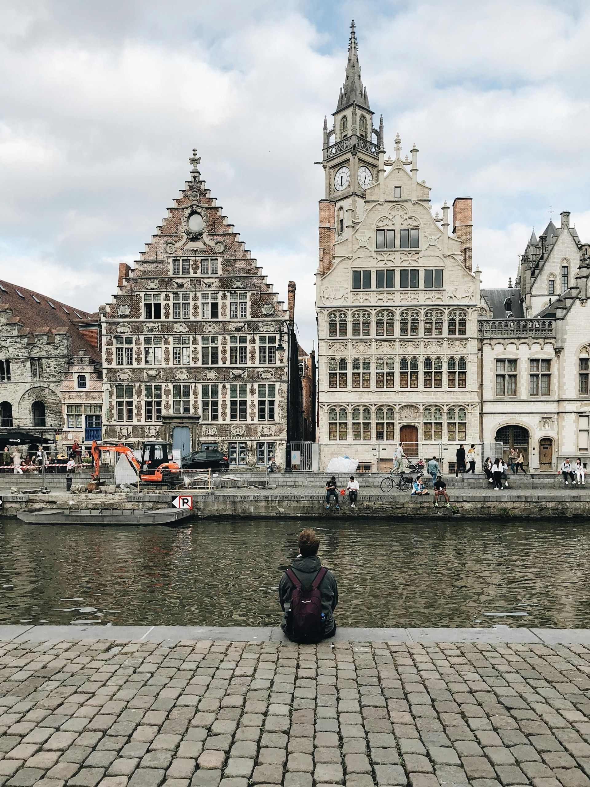 Une personne avec un sac à dos est assise sur un quai pavé, regardant de l'autre côté d'un canal des maisons historiques à pignons sous un ciel nuageux.