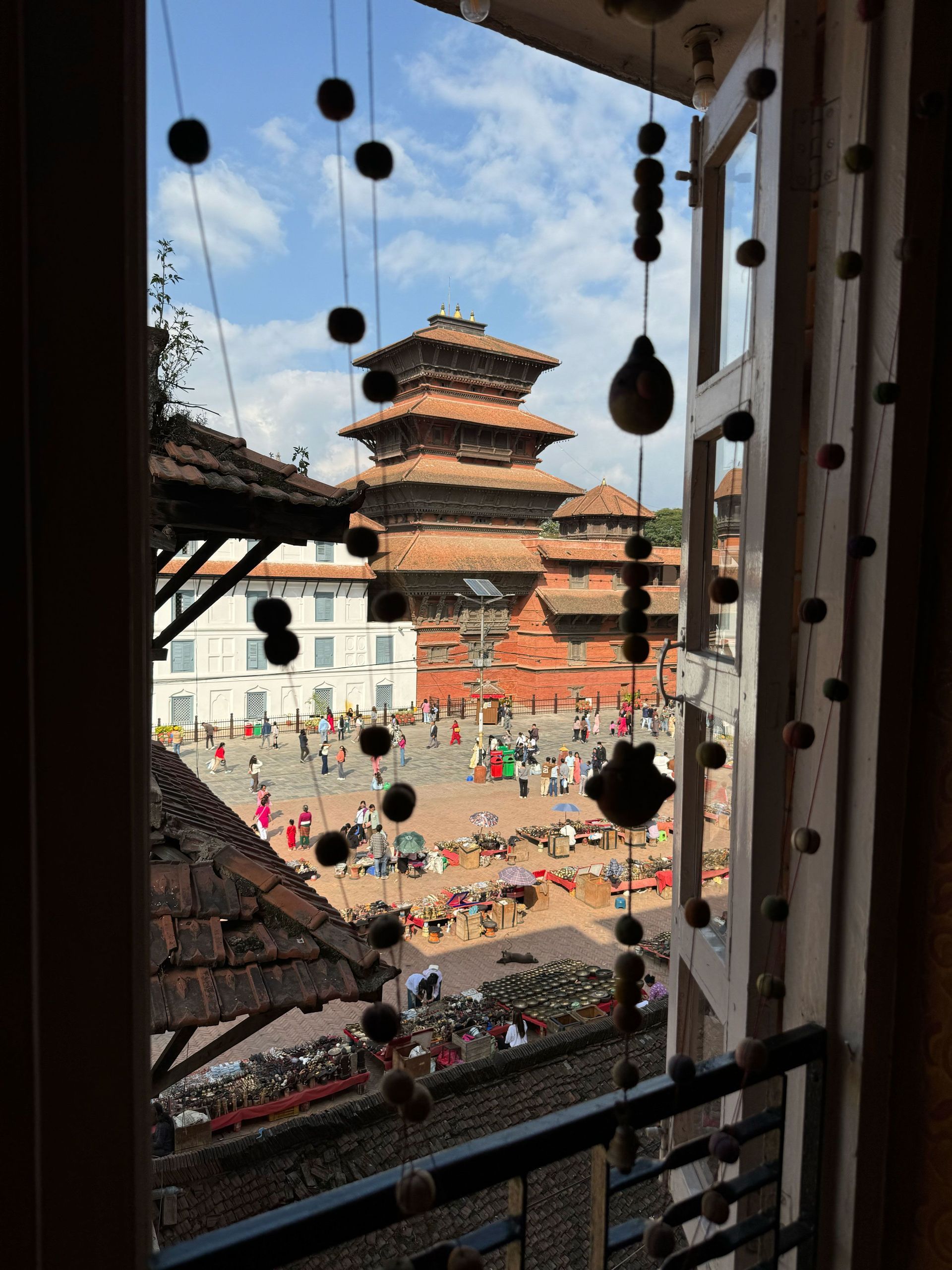 Una vista desde una ventana que da a una concurrida plaza pública con puestos de mercado, gente y un templo pagoda tradicional al fondo.
