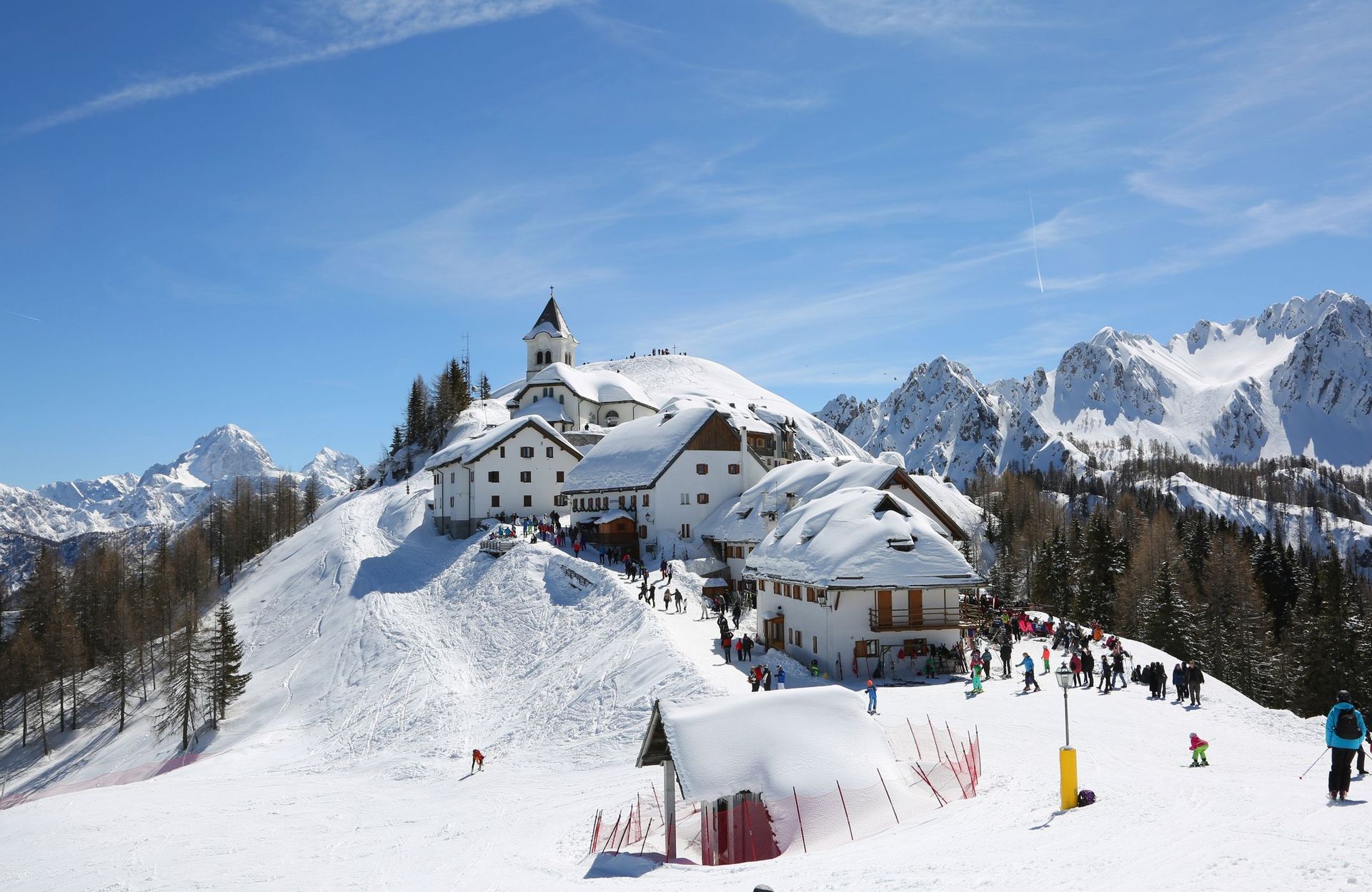 Un villaggio alpino innevato con una chiesa su una collina, circondato da sciatori e vette innevate sotto un cielo azzurro brillante.