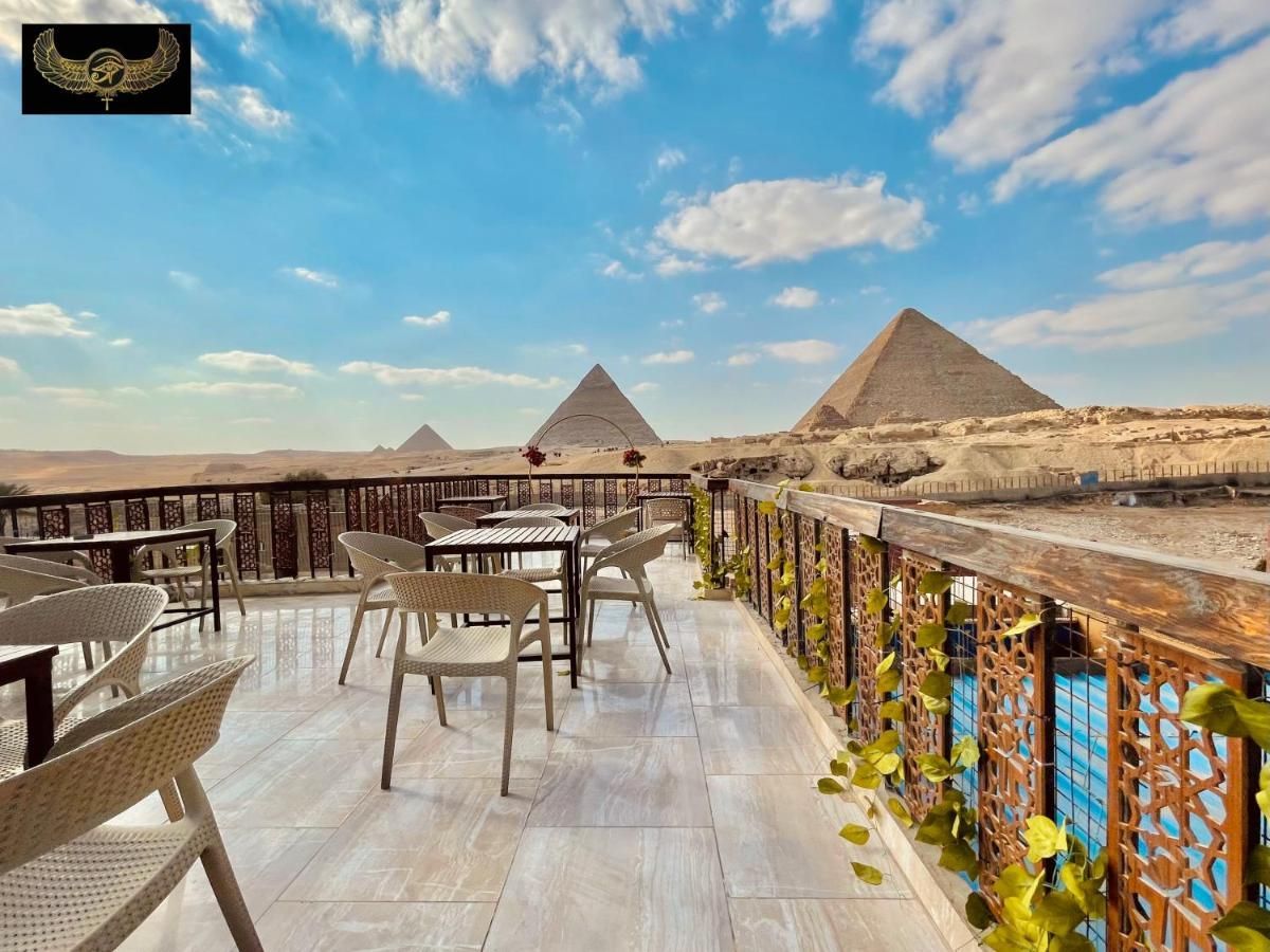 Una terrazza all'aperto con tavoli e sedie vuoti che si affaccia su tre grandi piramidi nel deserto sotto un cielo parzialmente nuvoloso.