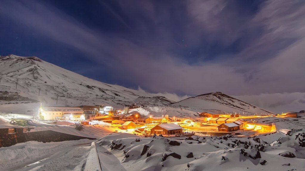 Un villaggio illuminato annidato tra montagne innevate di notte, con un cielo blu scuro e nuvole sopra.
