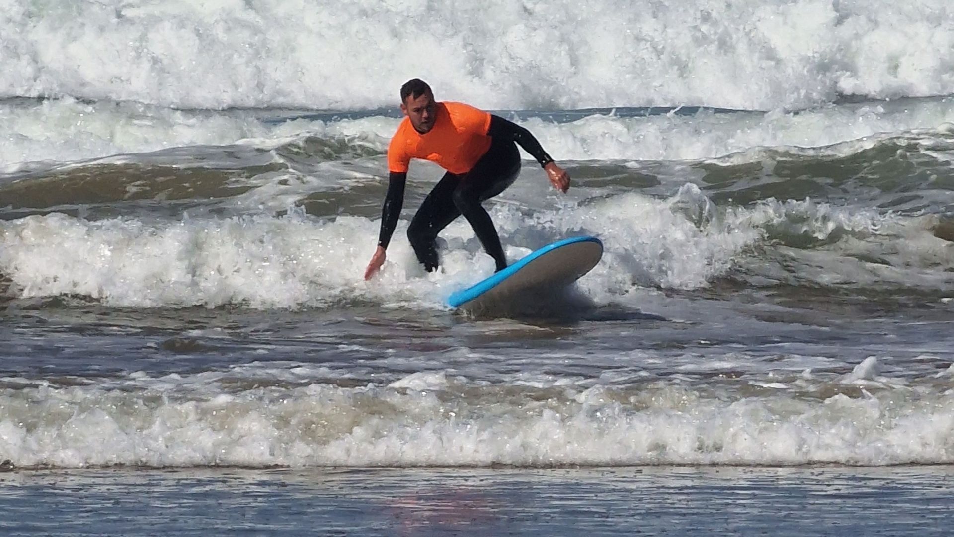 Ein Mann in einem orangefarbenen Hemd und schwarzem Neoprenanzug reitet eine Welle auf einem hellblauen Surfbrett.