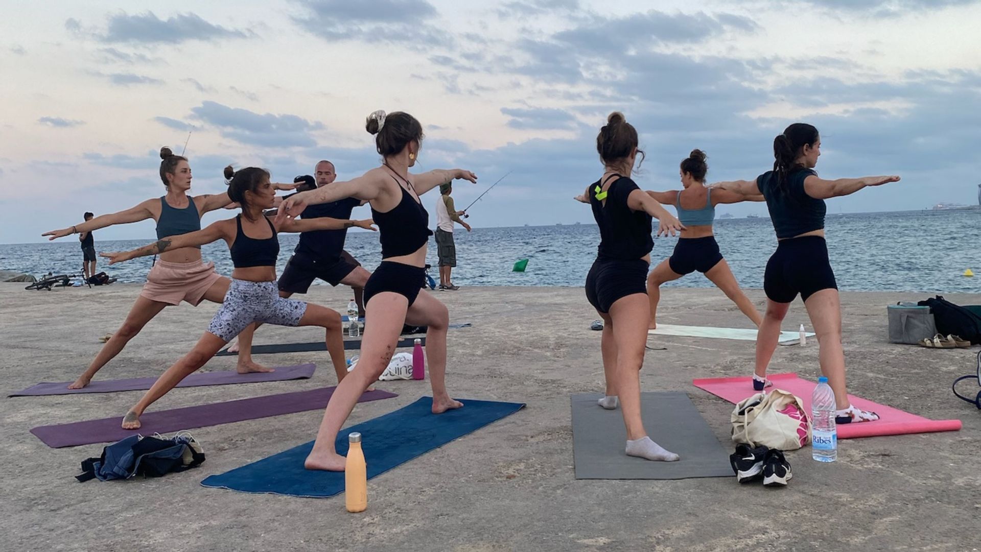 Eine WeRoad-Gruppenreise, die eine Yoga-Pose auf Matten auf einem Betonsteg am Meer unter bewölktem Himmel übt.