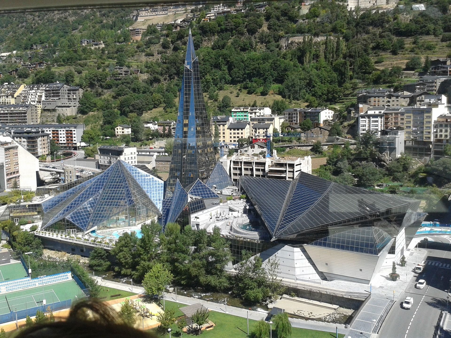 Veduta aerea di un moderno complesso architettonico con piramidi di vetro e un'alta guglia, situato in una città ai piedi di una montagna verde e boscosa.