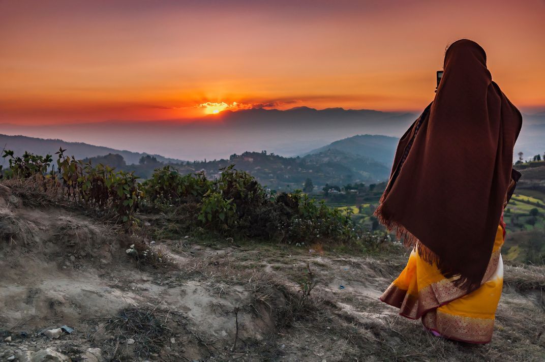Una donna in sari e scialle è su una collina, fotografando l'alba su una catena montuosa con il suo telefono.