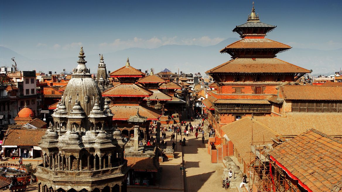 Una vista dall'alto di una vivace piazza cittadina con templi in pietra ornati e in stile pagoda, con montagne sullo sfondo.