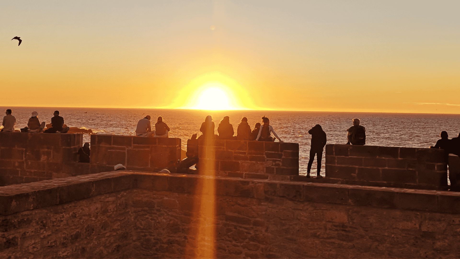 Silhouetten einer WeRoad-Gruppenreise, die den Sonnenuntergang über dem Meer von einer Steinmauer aus beobachten.