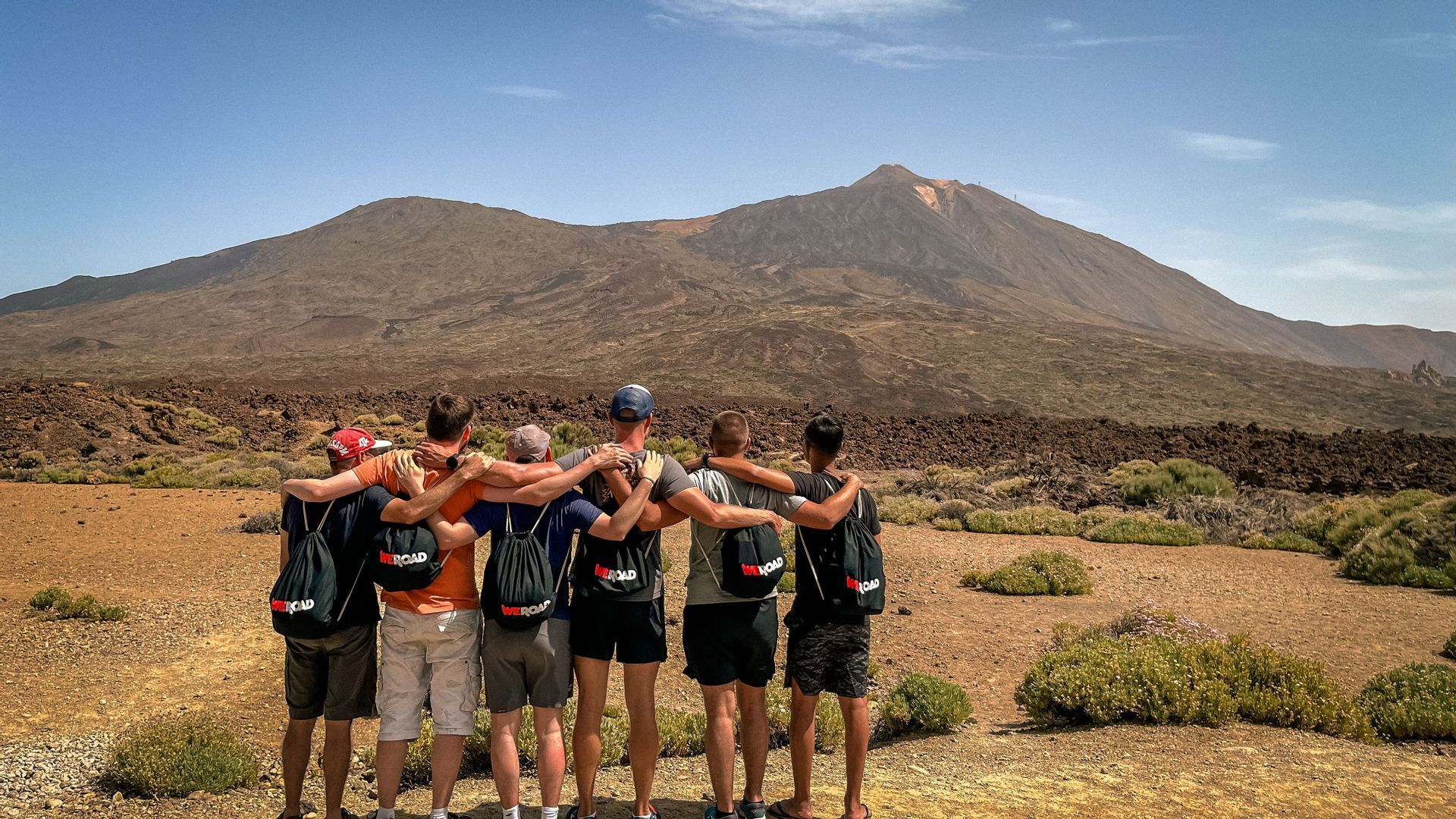 Sei uomini di un gruppo WeRoad si abbracciano, ammirando un vasto paesaggio montano vulcanico.