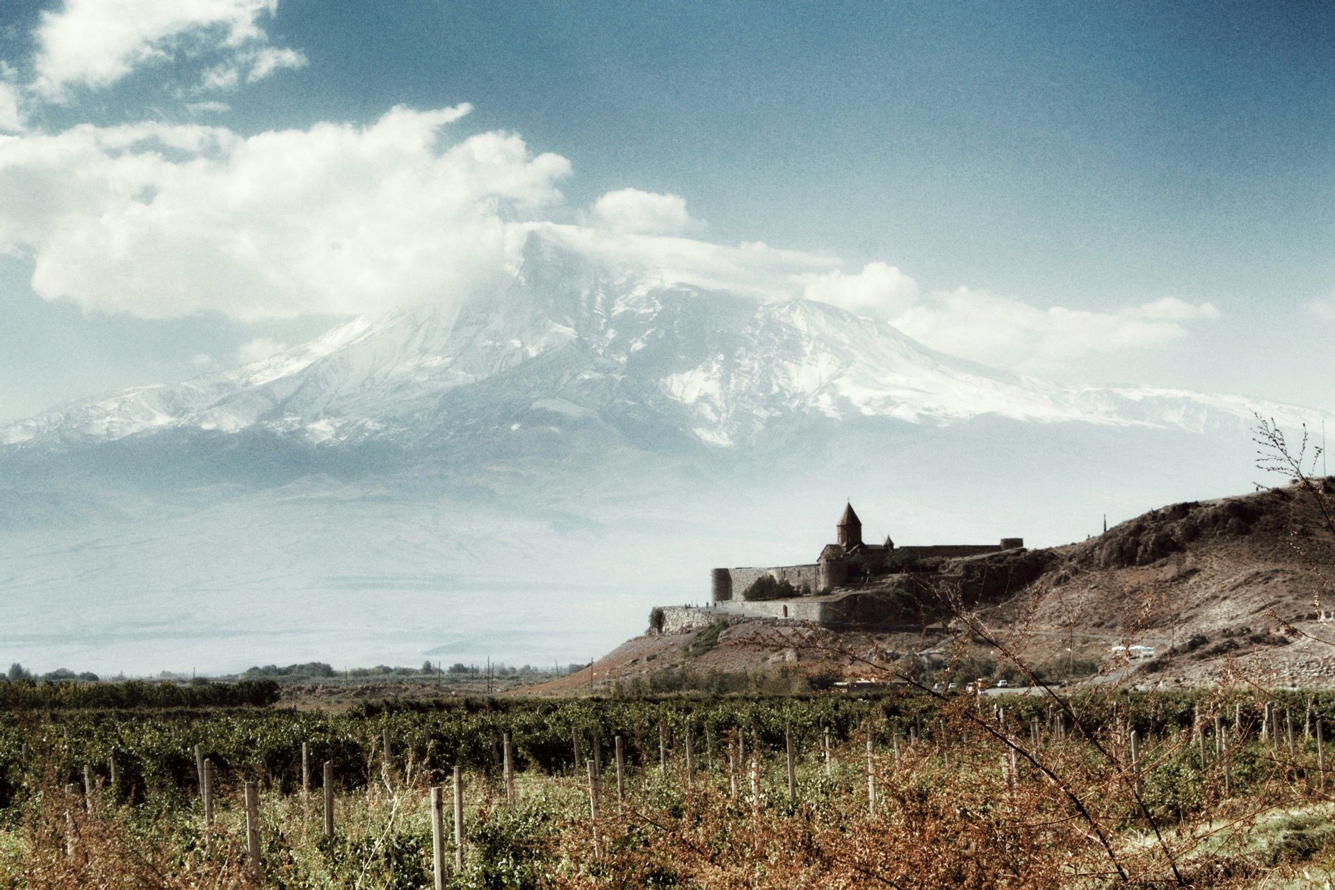 Un monastero in pietra si erge su una collina che domina un vasto vigneto, con una grande montagna innevata sullo sfondo sotto un cielo blu.