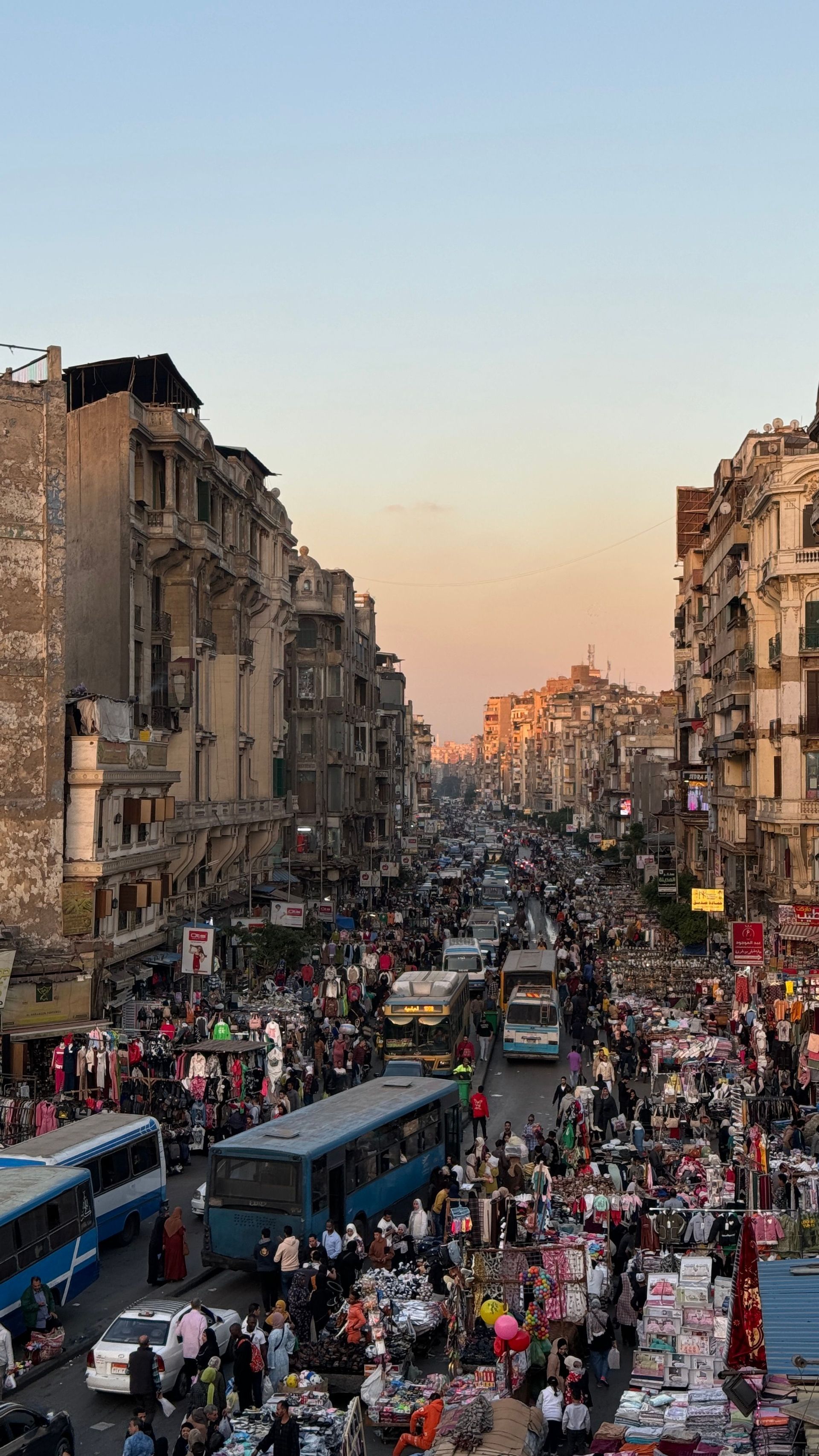Una veduta dall'alto di una vivace strada cittadina affollata di persone, bancarelle, autobus e automobili tra alti edifici al tramonto.