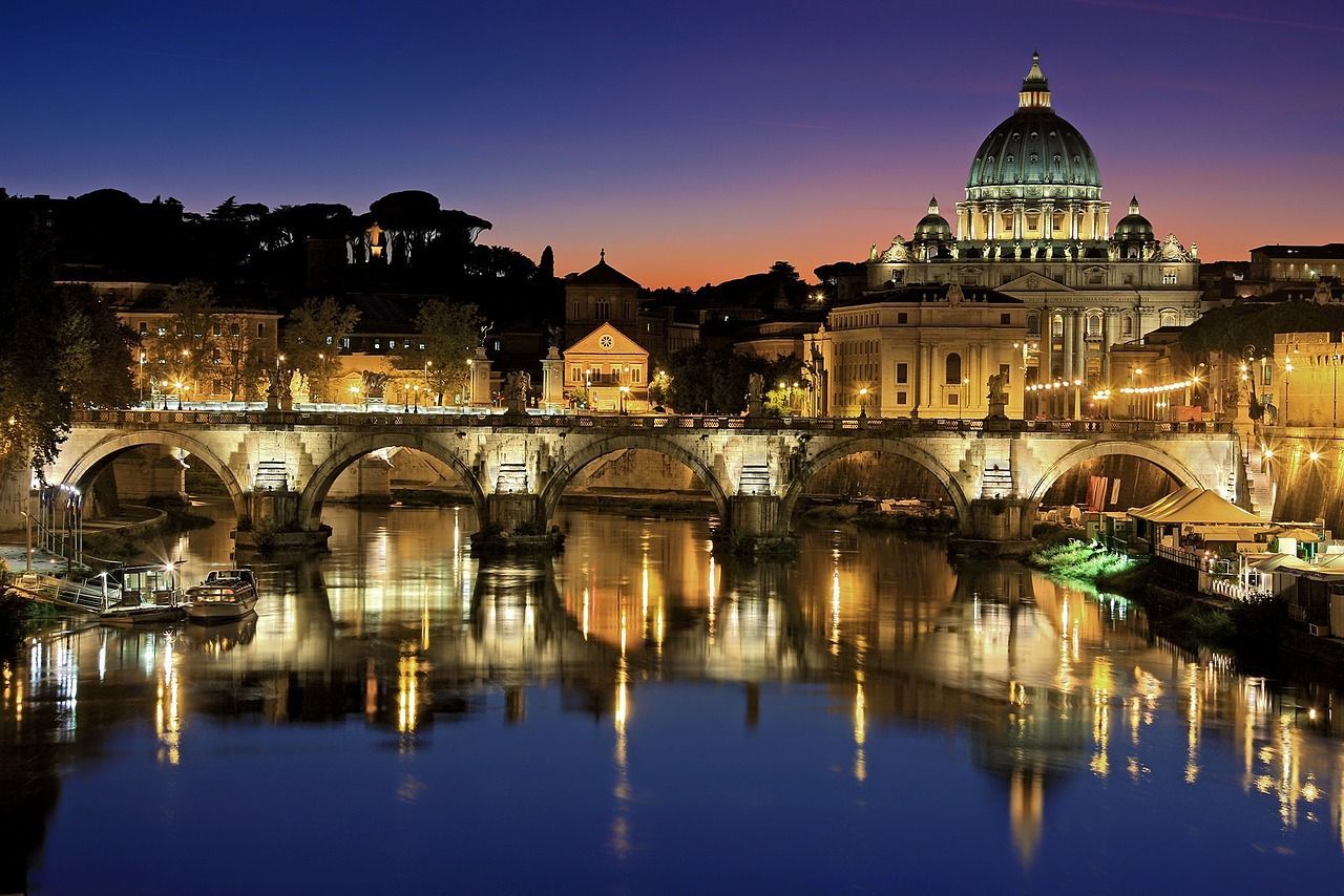 An illuminated stone bridge and a grand basilica at dusk, with their lights reflecting in the river below.