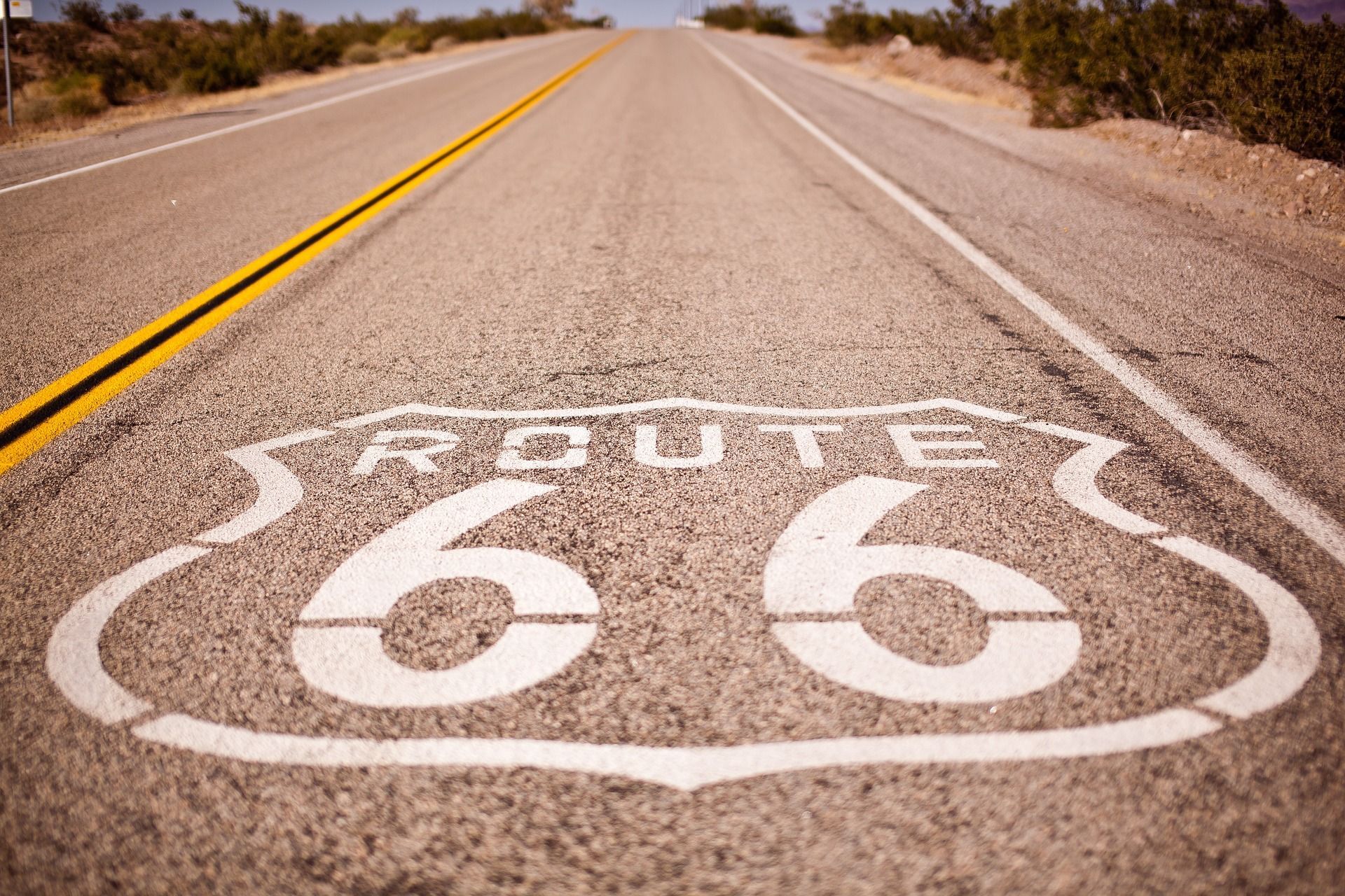 Un primer plano del cartel de la Ruta 66 pintado en una carretera de asfalto que se extiende a través de un paisaje seco y arbustivo.