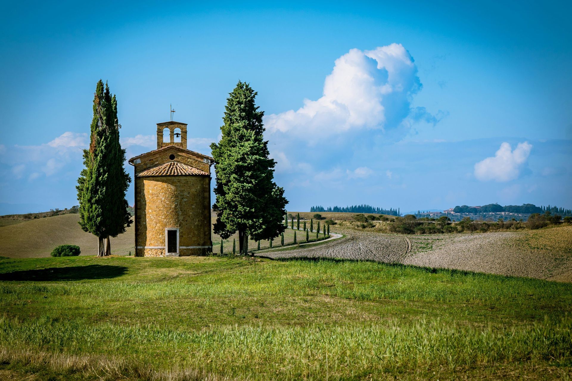 Una cappella in pietra con campanile si erge tra due cipressi su una collina verde sotto un cielo azzurro con nuvole.