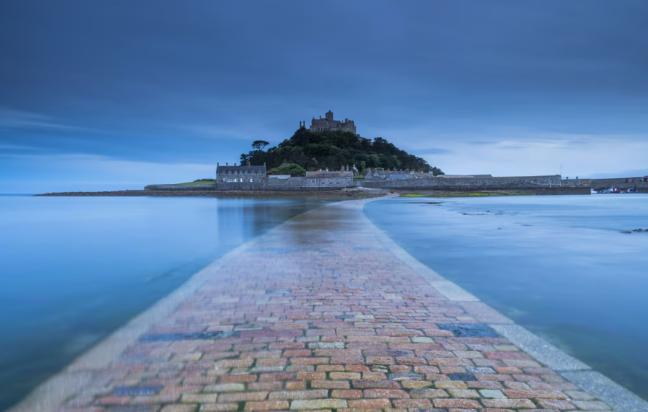Una strada lastricata di ciottoli conduce attraverso acque blu calme a un'isola con un castello sulla sua vetta sotto un cielo cupo.