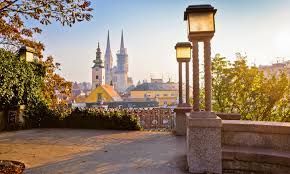 Vista da una terrazza in pietra con due lampade accese, affacciata su un panorama urbano con una cattedrale a due guglie al tramonto.