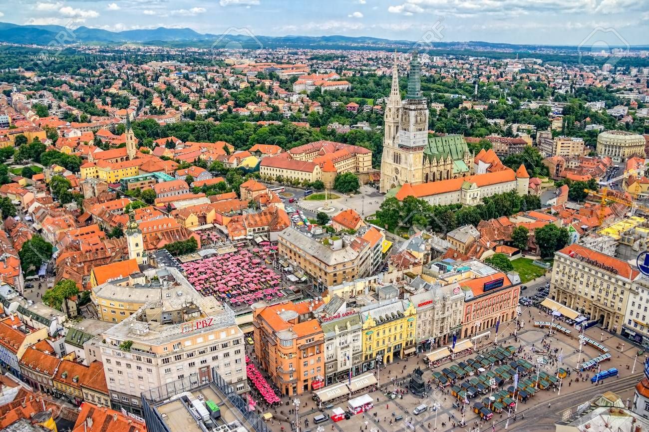 Vista aerea di una città europea con edifici dai tetti arancioni, una cattedrale a doppia guglia e una piazza del mercato con ombrelloni rossi.