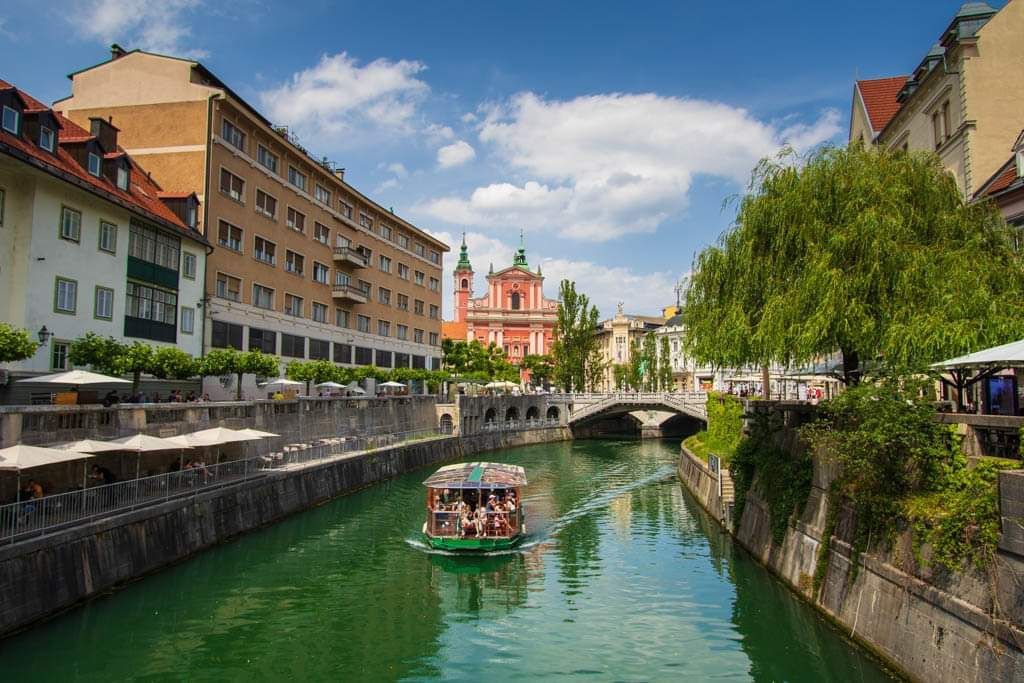 Una barca turistica naviga lungo un canale cittadino costeggiato da edifici storici, un ponte in pietra e un grande salice piangente sotto un cielo azzurro.