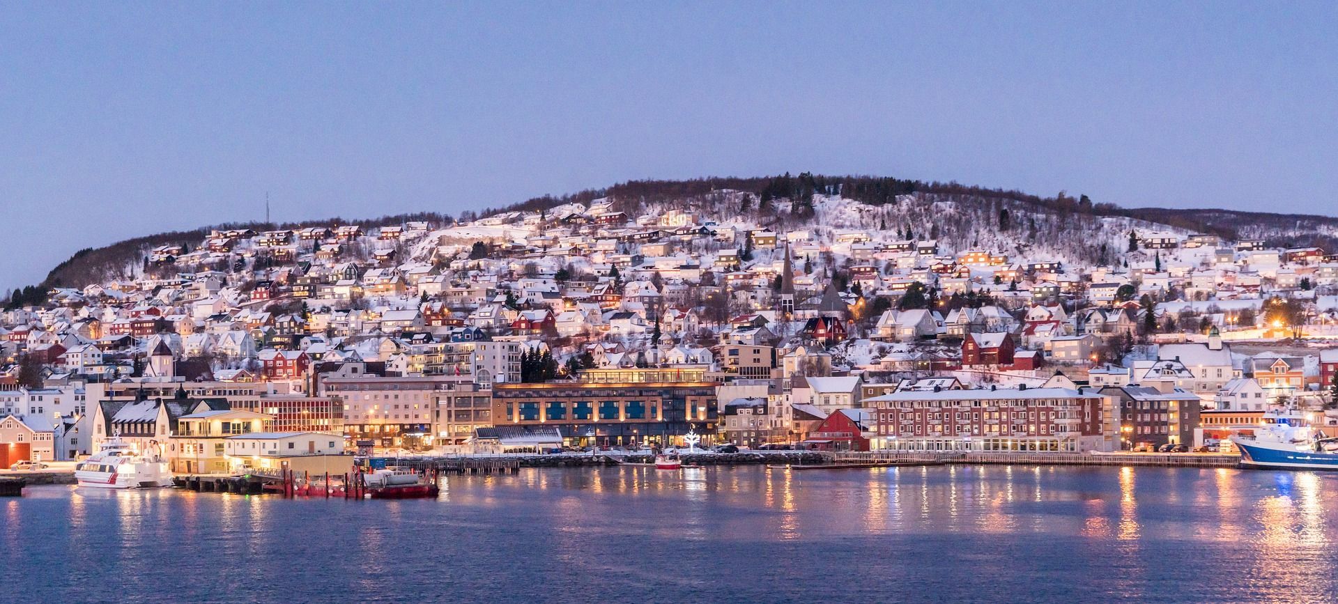 A snow-covered town built on a hillside along the water, with its buildings illuminated at dusk.
