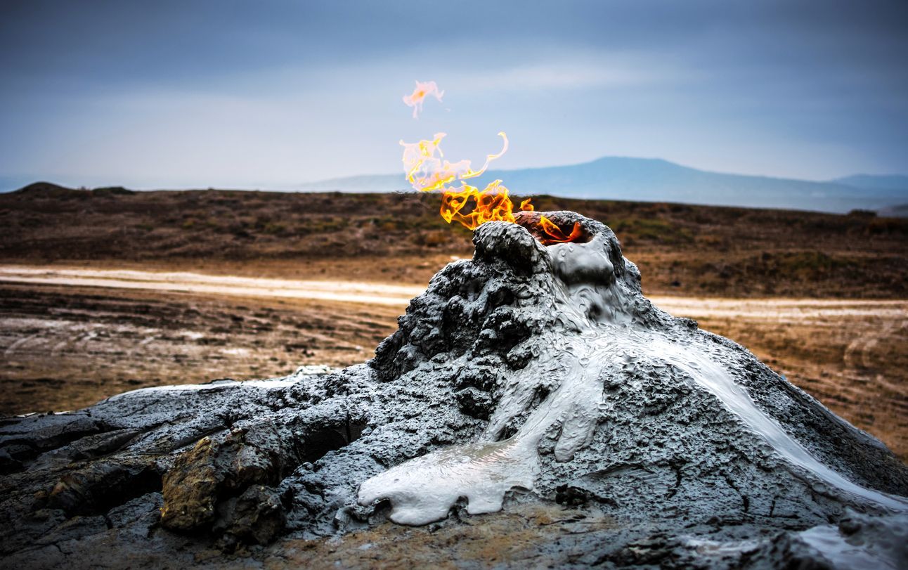 Un piccolo vulcano di fango erutta fiamme arancioni brillanti dal suo cratere in un paesaggio brullo sotto un cielo coperto.