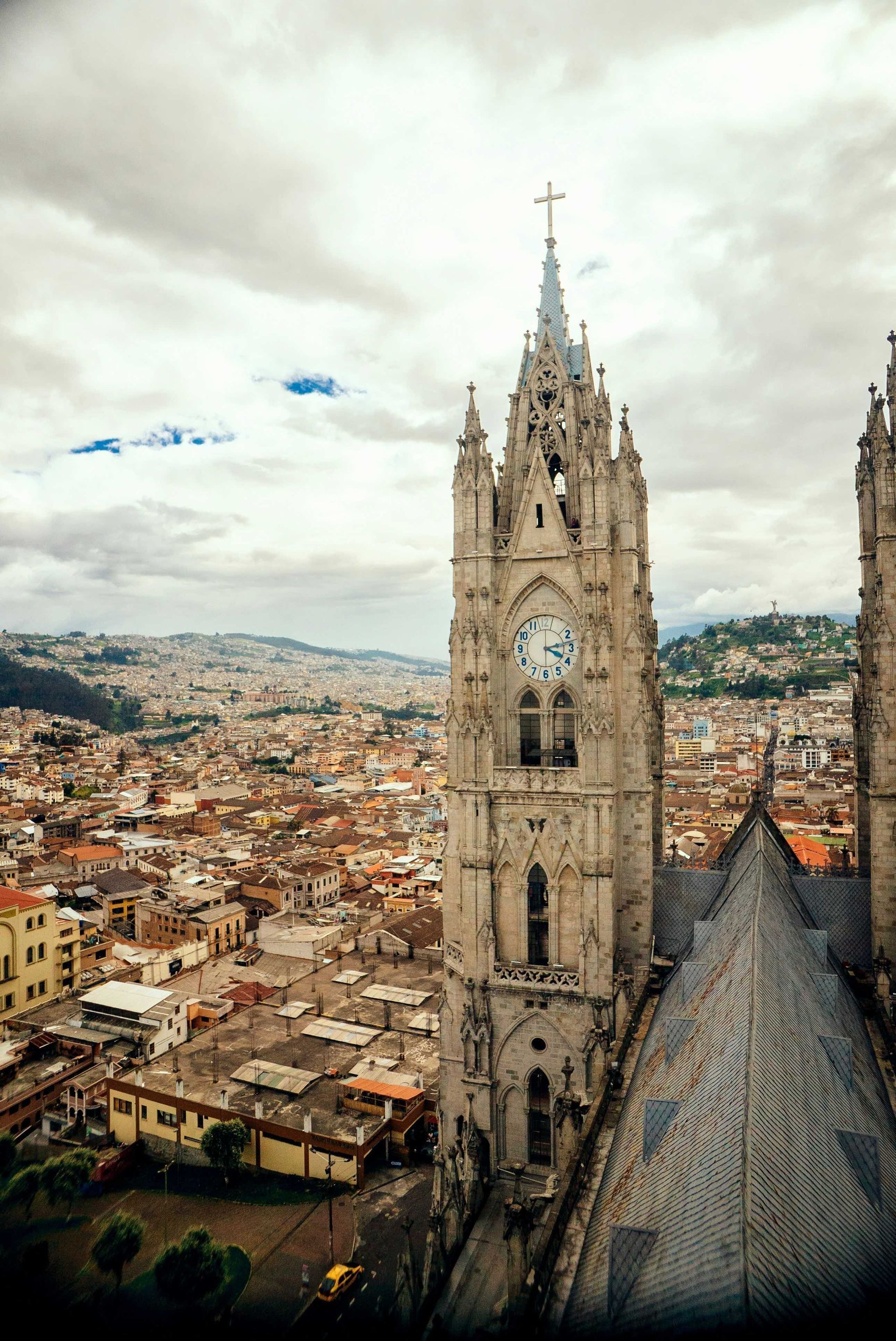 A tall, Gothic cathedral clock tower overlooking a cityscape that extends to distant hills under a cloudy sky.
