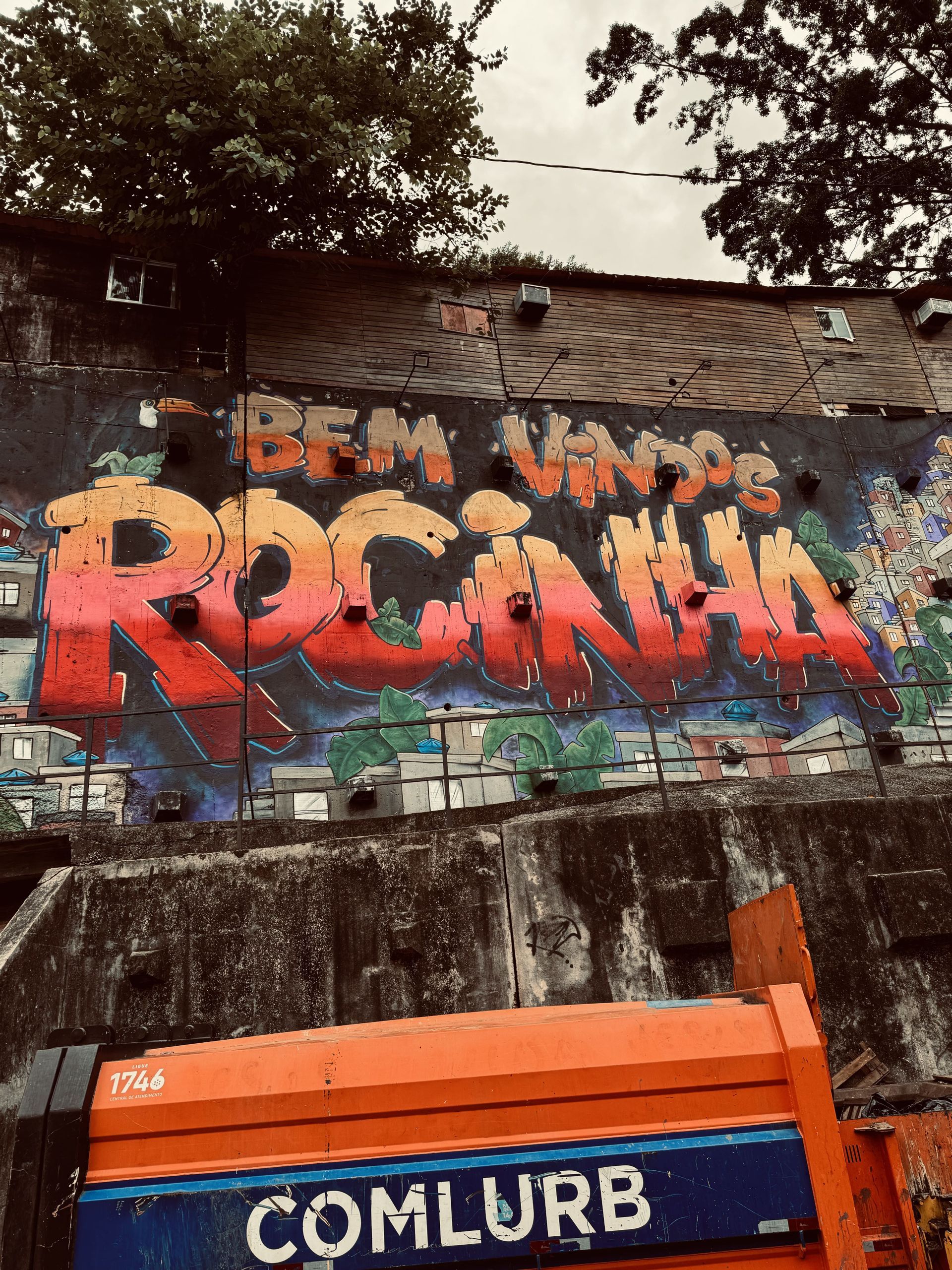 A colorful graffiti mural that says 'Bem Vindos Rocinha' on a concrete wall, with an orange municipal truck in the foreground.