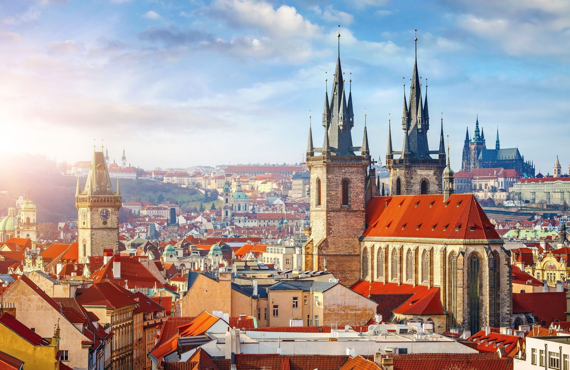 Vista aérea del horizonte de una ciudad histórica con agujas góticas, una torre del reloj y tejados rojos bajo un cielo soleado.