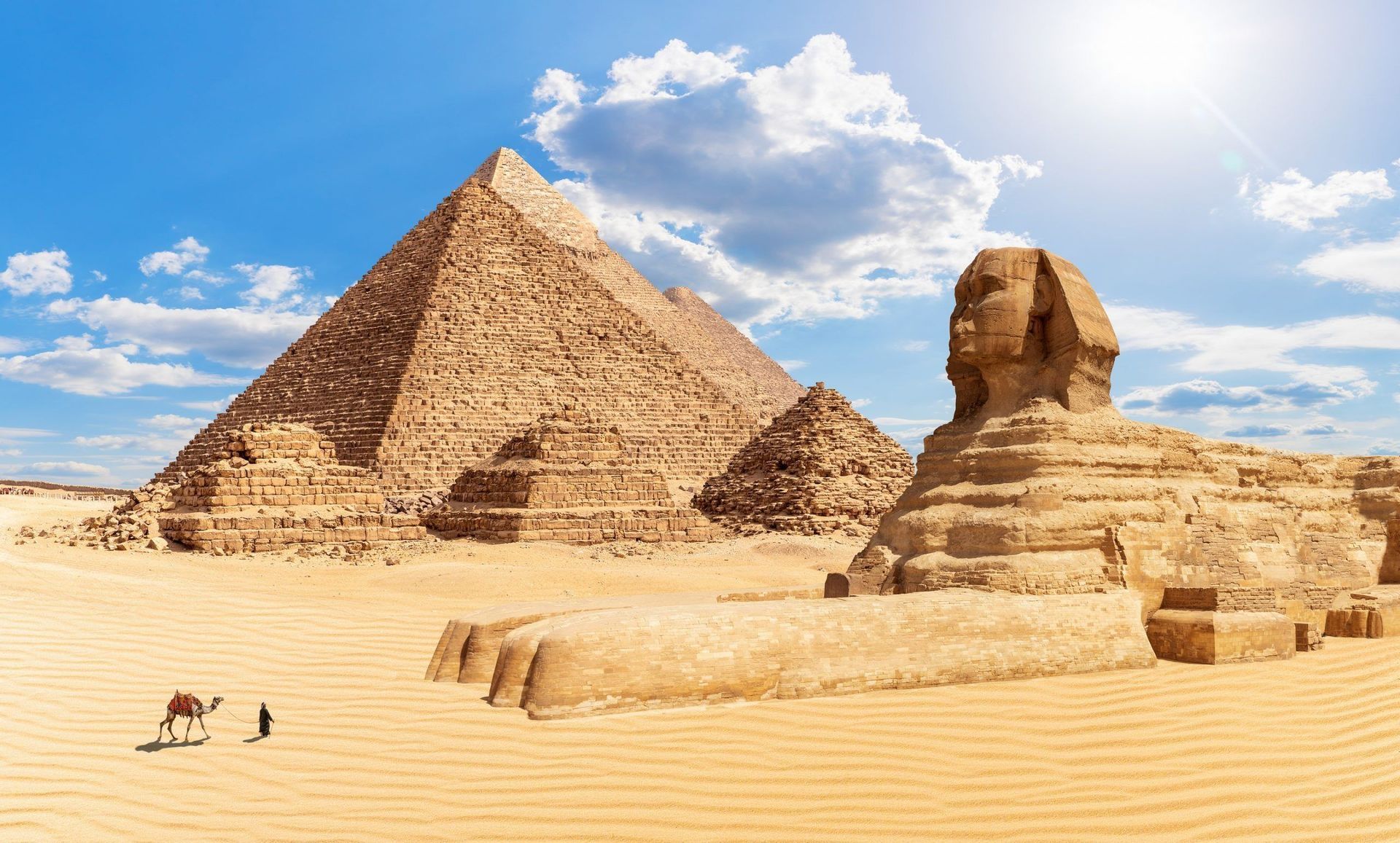 La Grande Sfinge e tre grandi piramidi si ergono nel deserto sotto un cielo azzurro e soleggiato, con una persona e un cammello in primo piano.