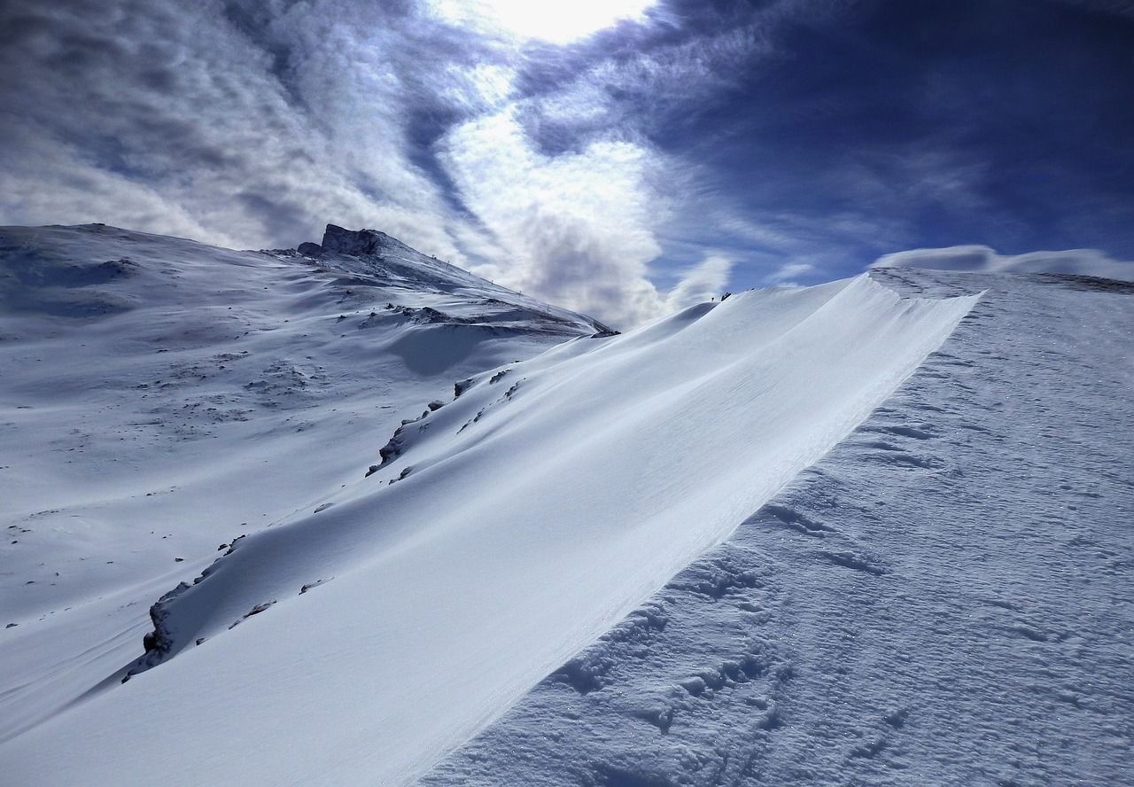Un paisaje montañoso cubierto de nieve con una cresta barrida por el viento bajo un cielo azul profundo con nubes arremolinadas.