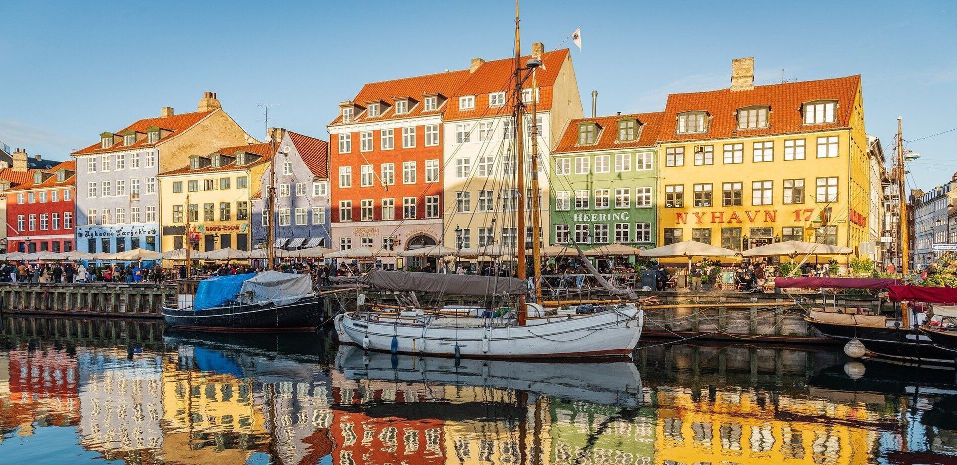 Barche a vela ormeggiate in un canale di fronte a edifici colorati sul lungomare, con i loro riflessi che brillano sull'acqua.