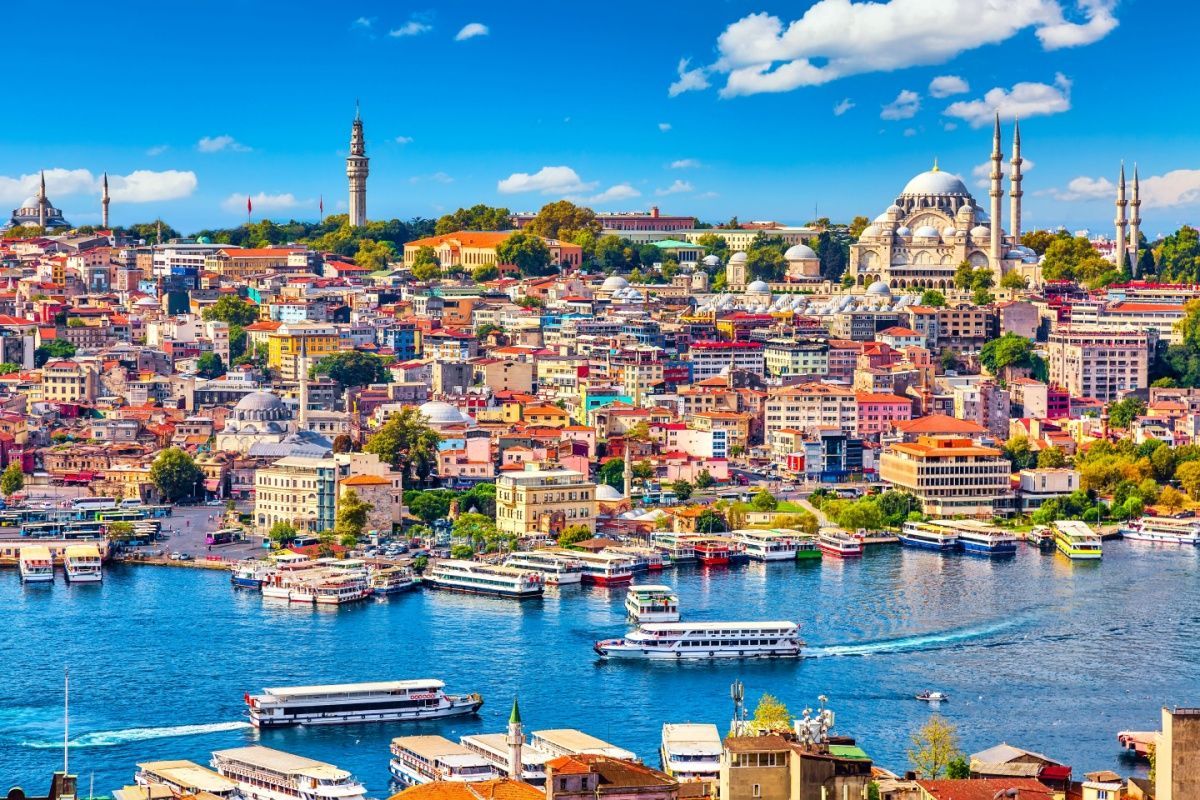 Una vista panoramica di una densa città costiera con moschee storiche e una torre, mentre le barche navigano sull'acqua blu sottostante.