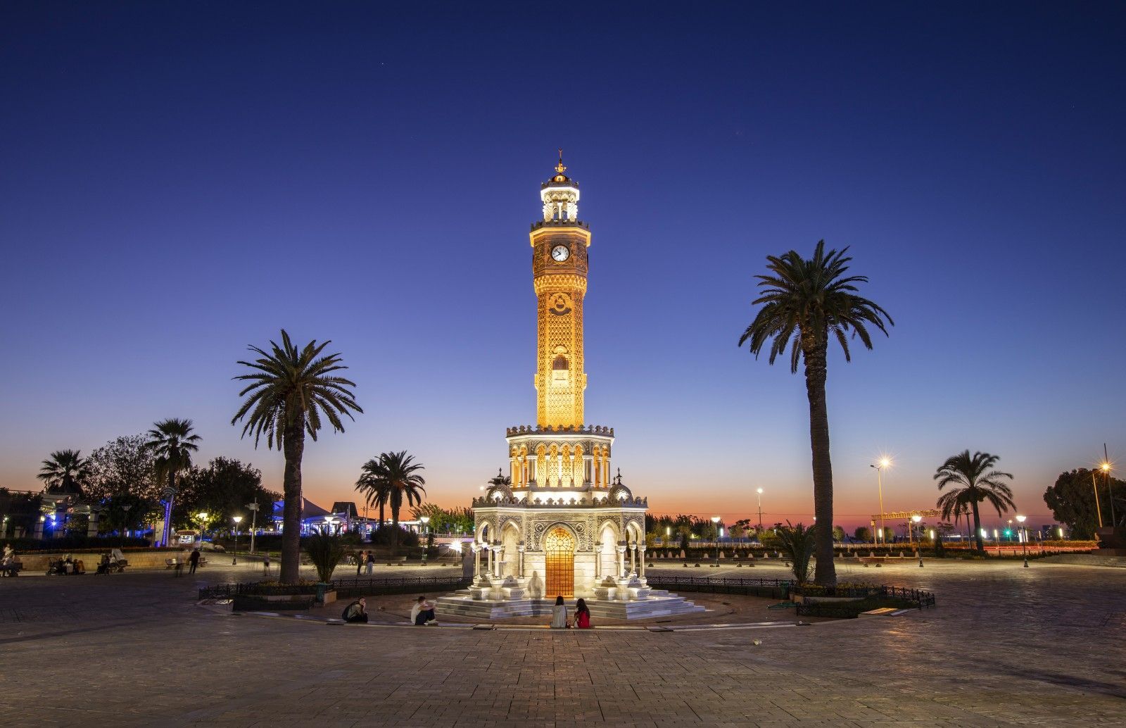 Una torre dell'orologio decorata si erge illuminata in una piazza pubblica al crepuscolo, circondata da alte palme.
