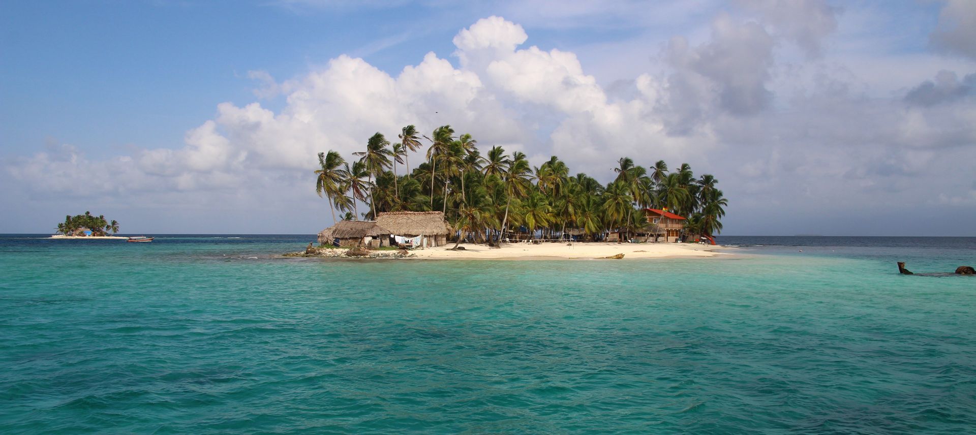 A small tropical island with palm trees and huts on a white sand beach, surrounded by turquoise water under a cloudy sky.