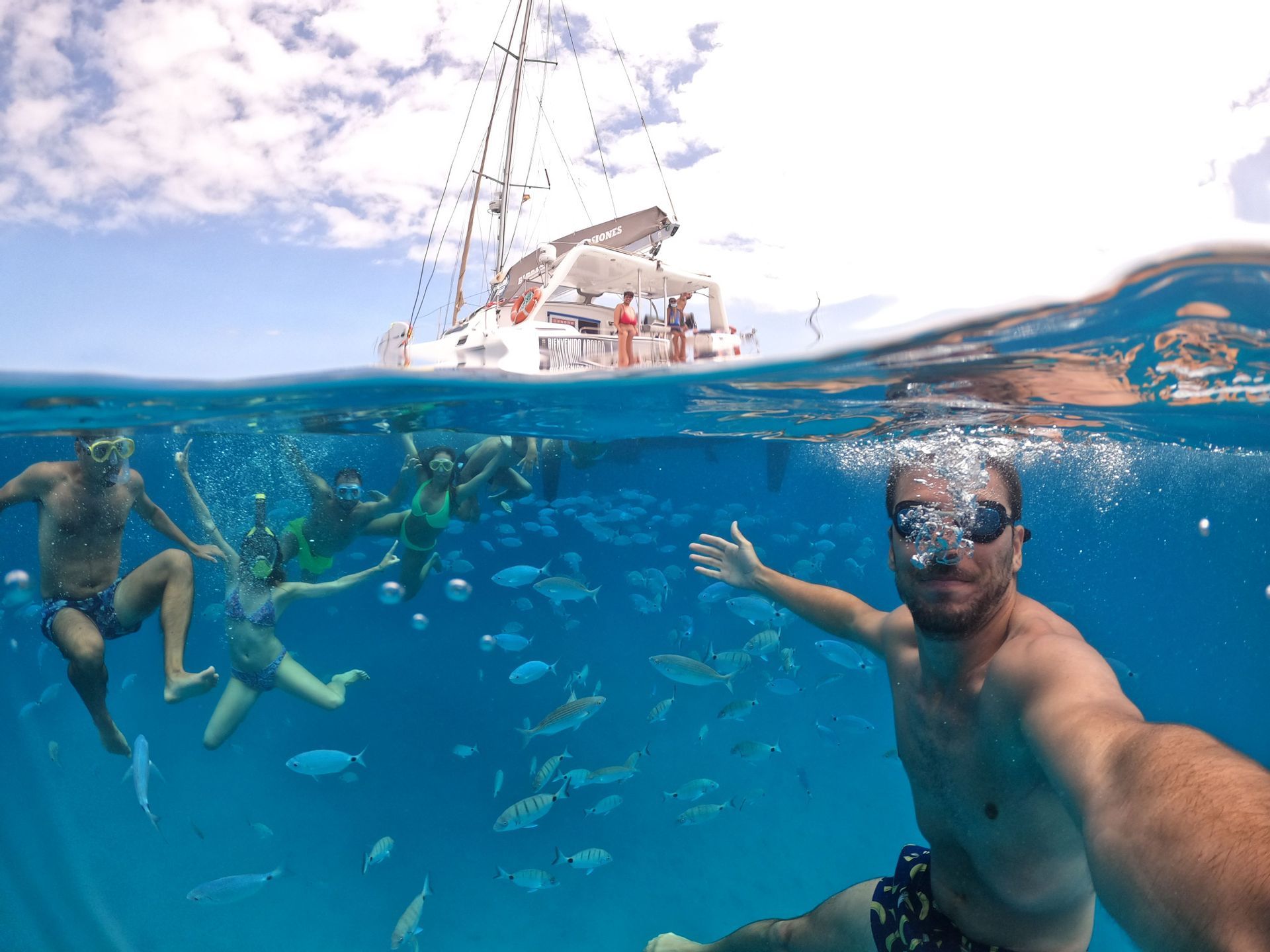 Un uomo si scatta un selfie subacqueo con il suo gruppo WeRoad mentre fa snorkeling tra un banco di pesci, con un catamarano in superficie.