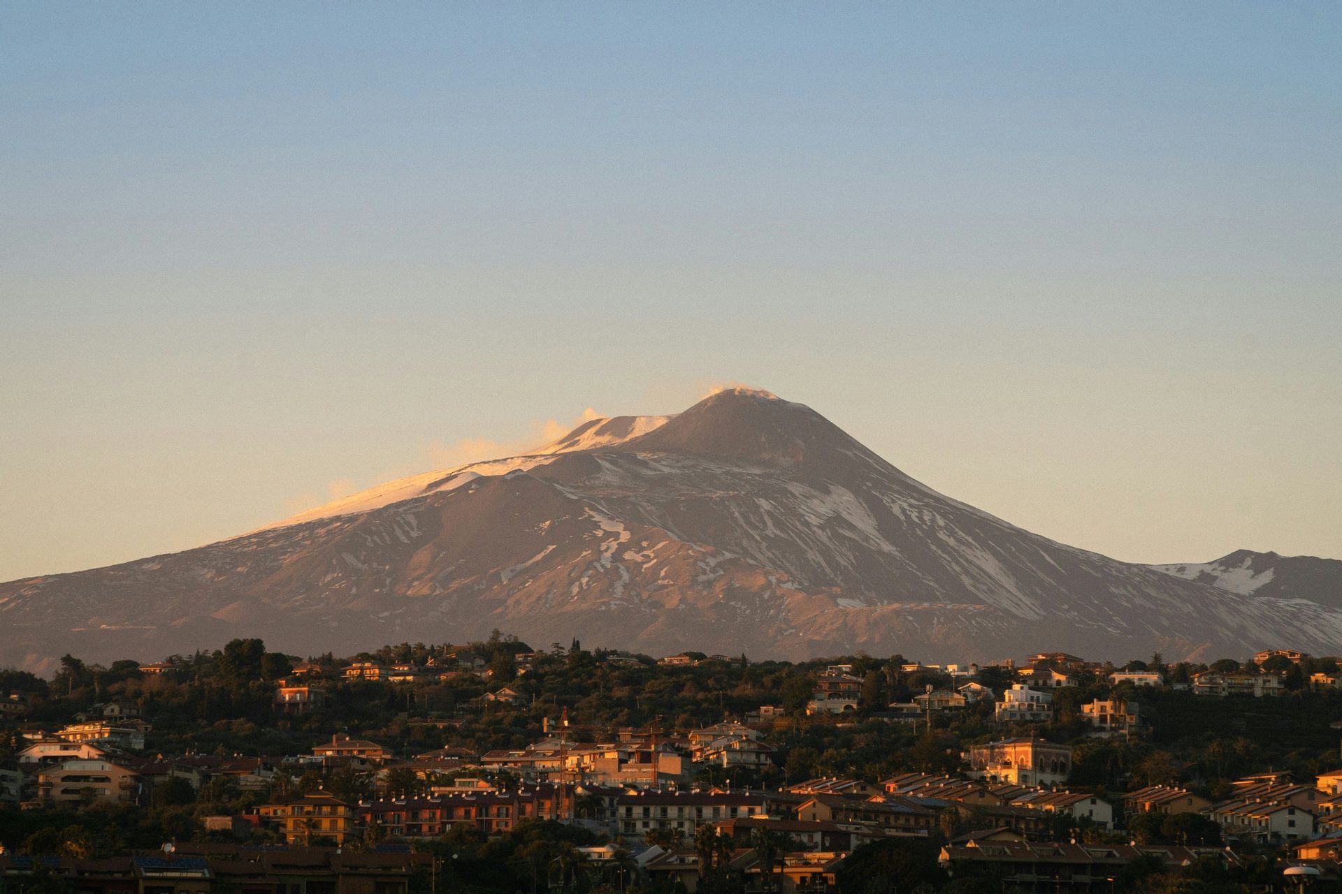 Un grande vulcano innevato, con un pennacchio di fumo dalla sua cima, si erge maestoso su una cittadina collinare bagnata dalla calda luce dell'alba.