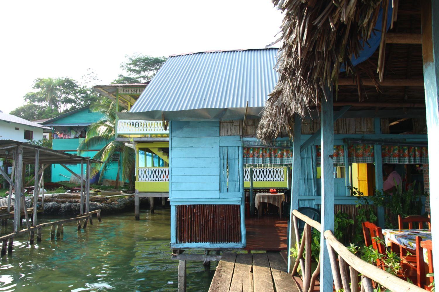 Des maisons colorées en bois sur pilotis se dressent sur l'eau dans un village tropical avec des passerelles en bois.
