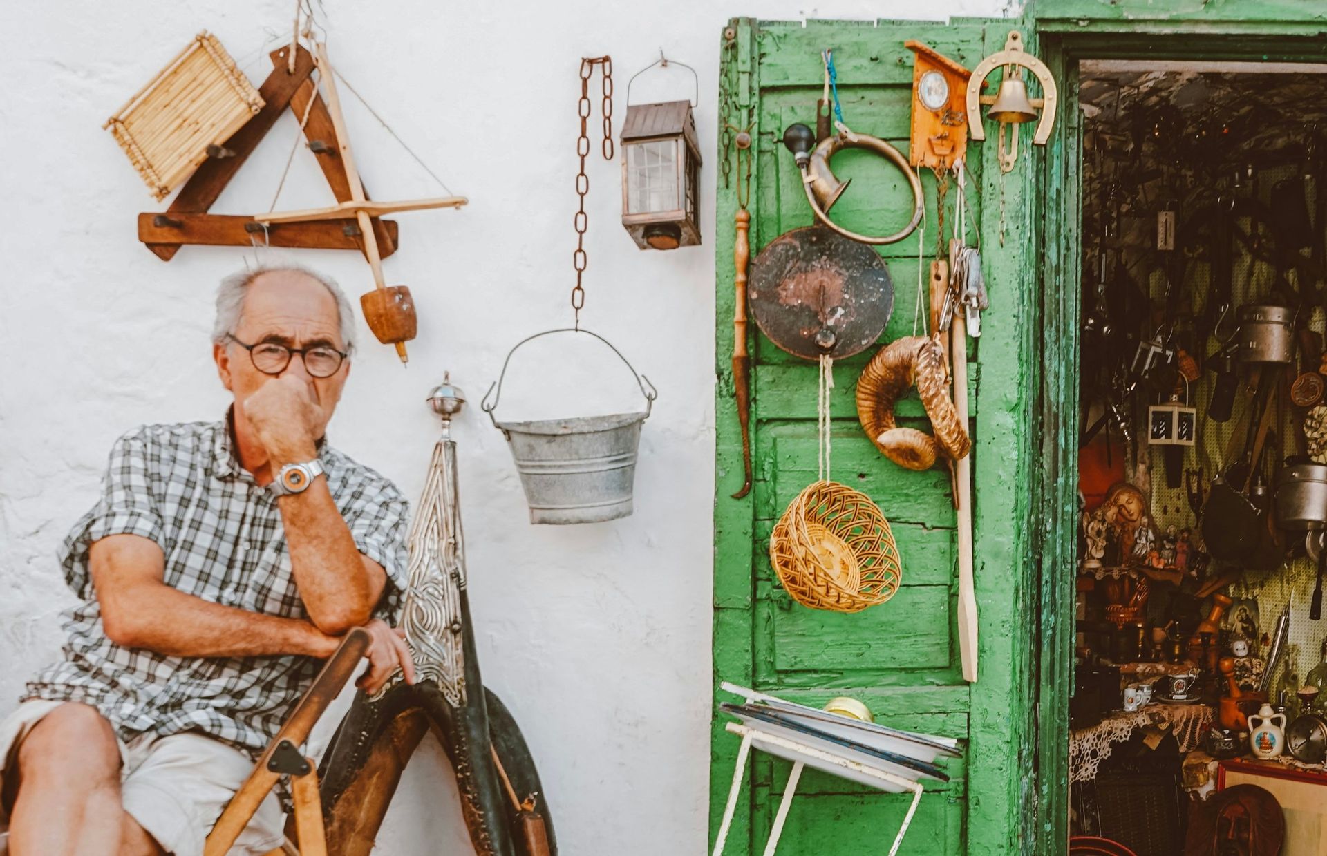 Un anziano signore siede pensieroso fuori da un negozio, la cui parete bianca e la porta verde sono decorate con vari oggetti vintage.