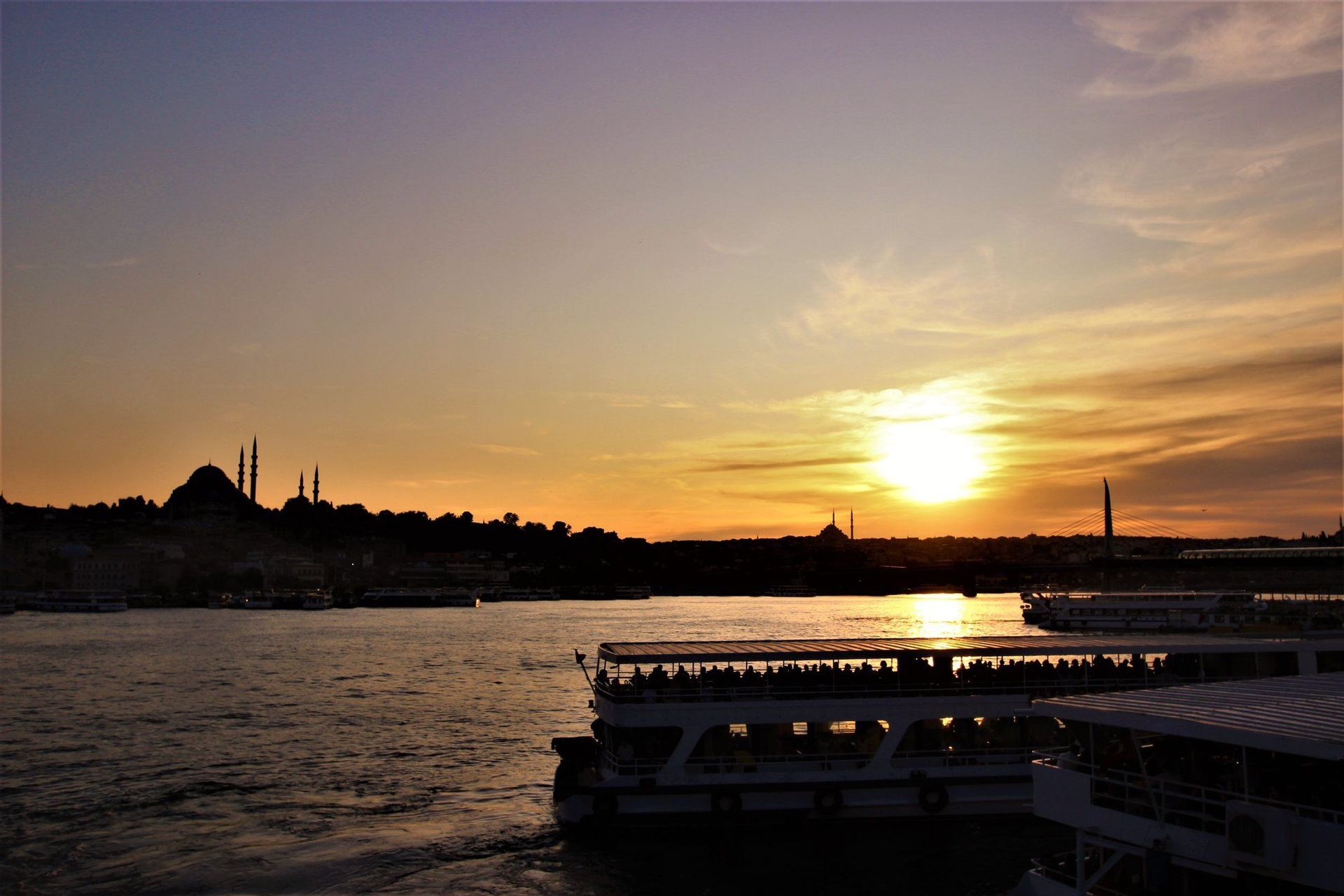Ferries con pasajeros navegan sobre el agua al atardecer, con el perfil de la ciudad y mezquitas en silueta al fondo.