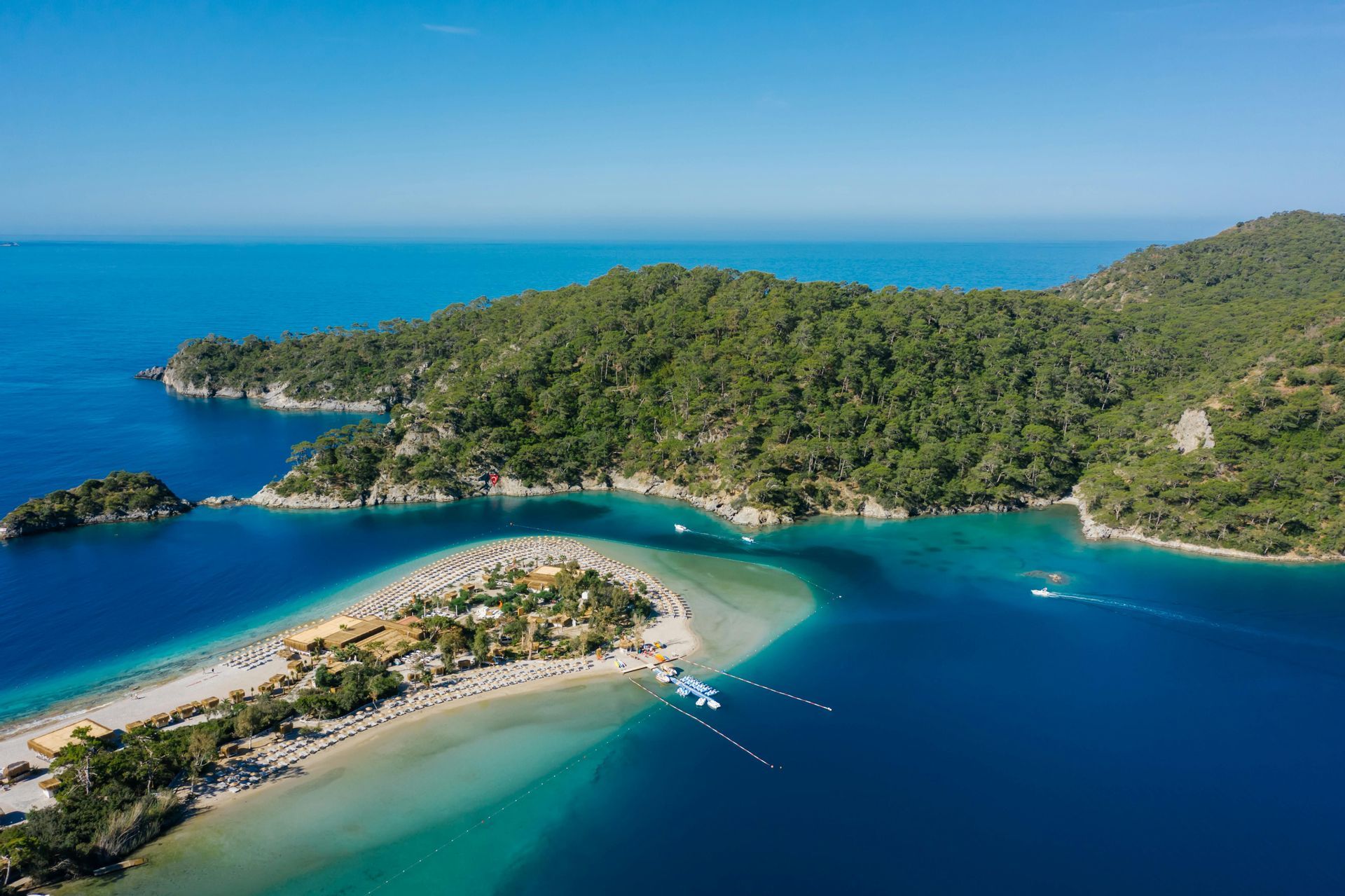 Vista aerea di una lingua di sabbia con ombrelloni, circondata da acque turchesi e blu intenso, vicino a una costa alberata.