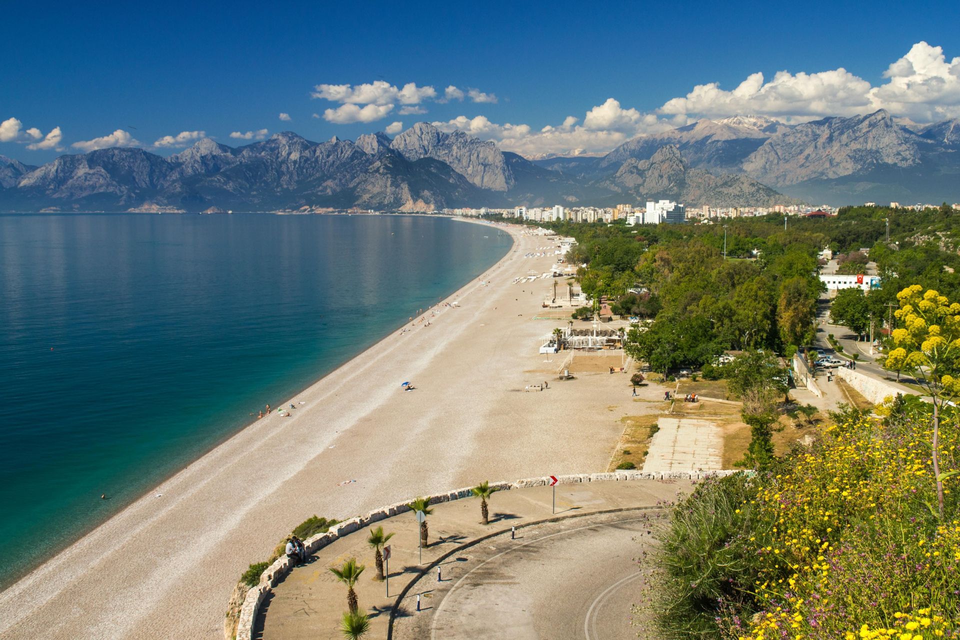 Una lunga spiaggia sabbiosa e curvilinea incontra il mare turchese, con una città costiera e un'imponente catena montuosa sullo sfondo.