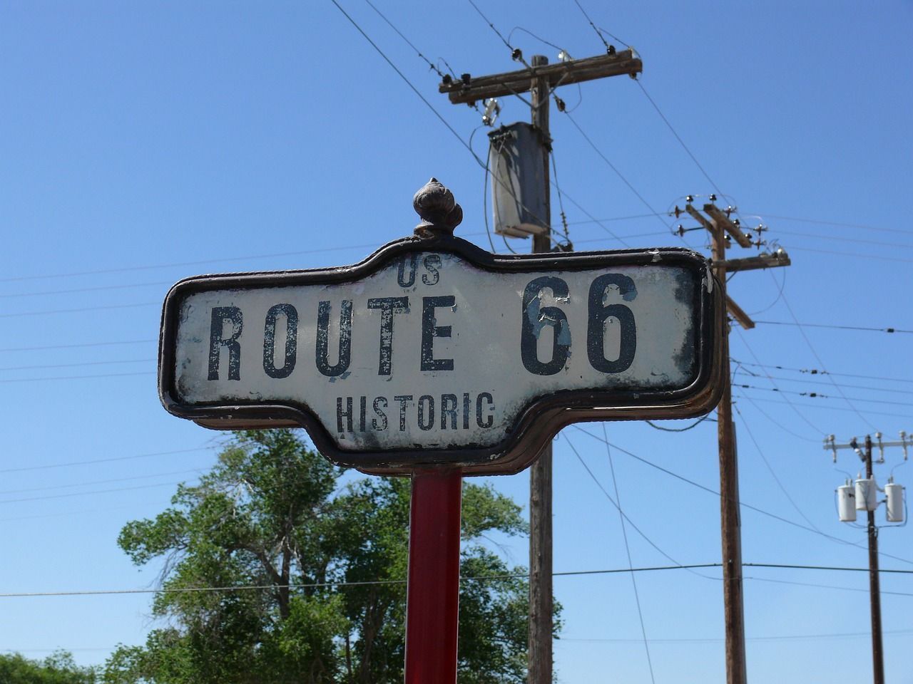 Primer plano de un cartel desgastado de la Ruta Histórica 66 de EE. UU. en un poste rojo, con un cielo azul y postes de servicios de fondo.