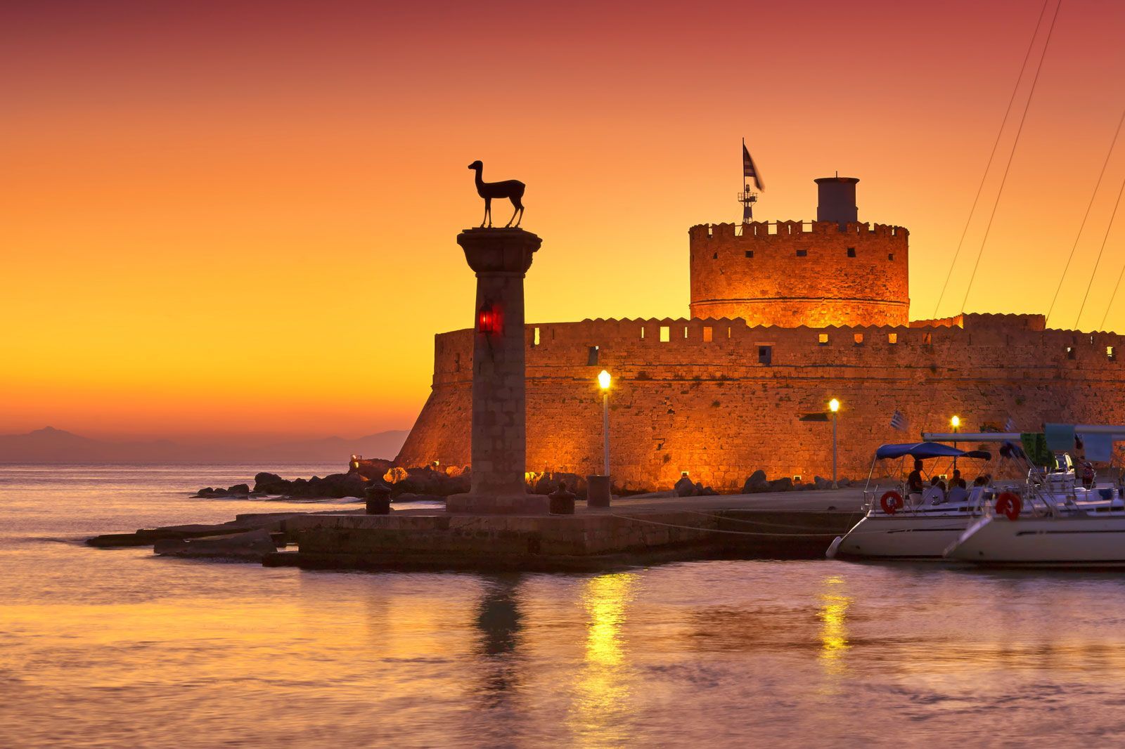 Un forte in pietra e un pilastro sormontato da una statua di cervo sono stagliati all'ingresso di un porto contro un tramonto arancione.
