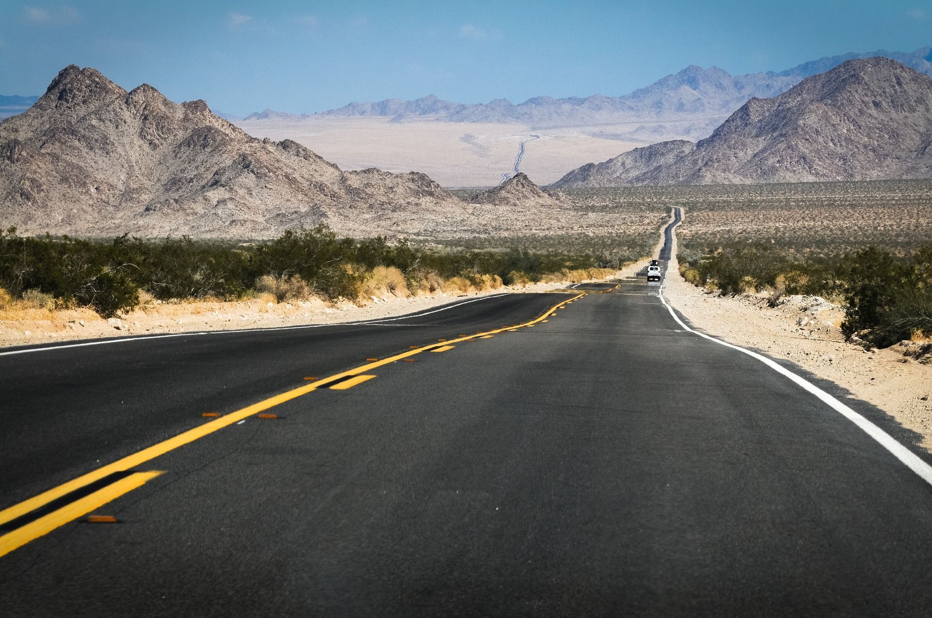 Una carretera pavimentada con líneas amarillas se extiende por un valle desértico, con montañas rocosas visibles a lo lejos bajo un cielo azul.