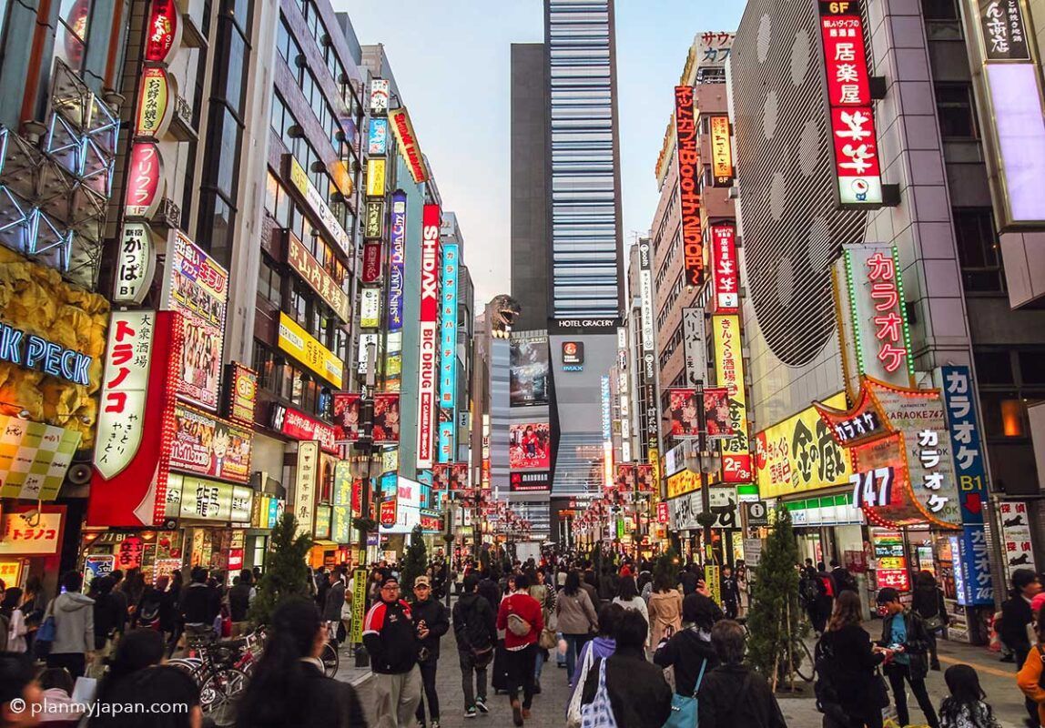 Una folla di persone cammina lungo una strada cittadina fiancheggiata da edifici coperti da colorate insegne al neon, con una grande testa di Godzilla visibile.