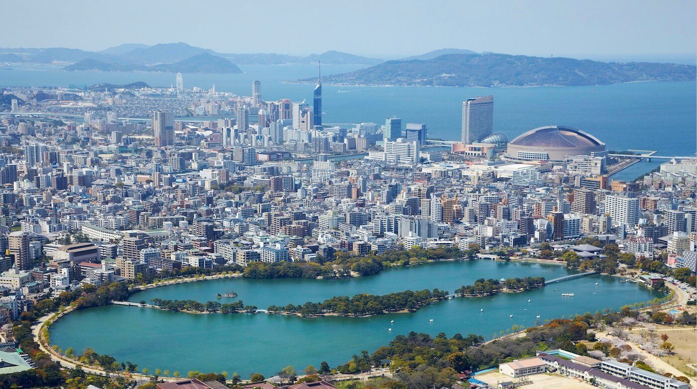Una vista aerea di una vasta città costiera con un grande lago serpeggiante in un parco, moderni grattacieli e uno stadio a cupola sul mare.